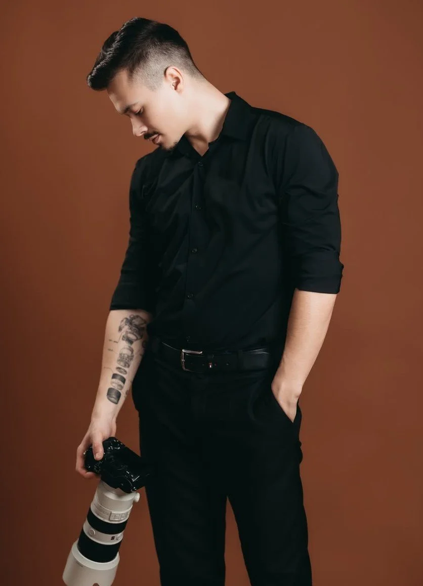 Young man in black shirt holding a professional camera with a large telephoto lens, standing against a brown background.