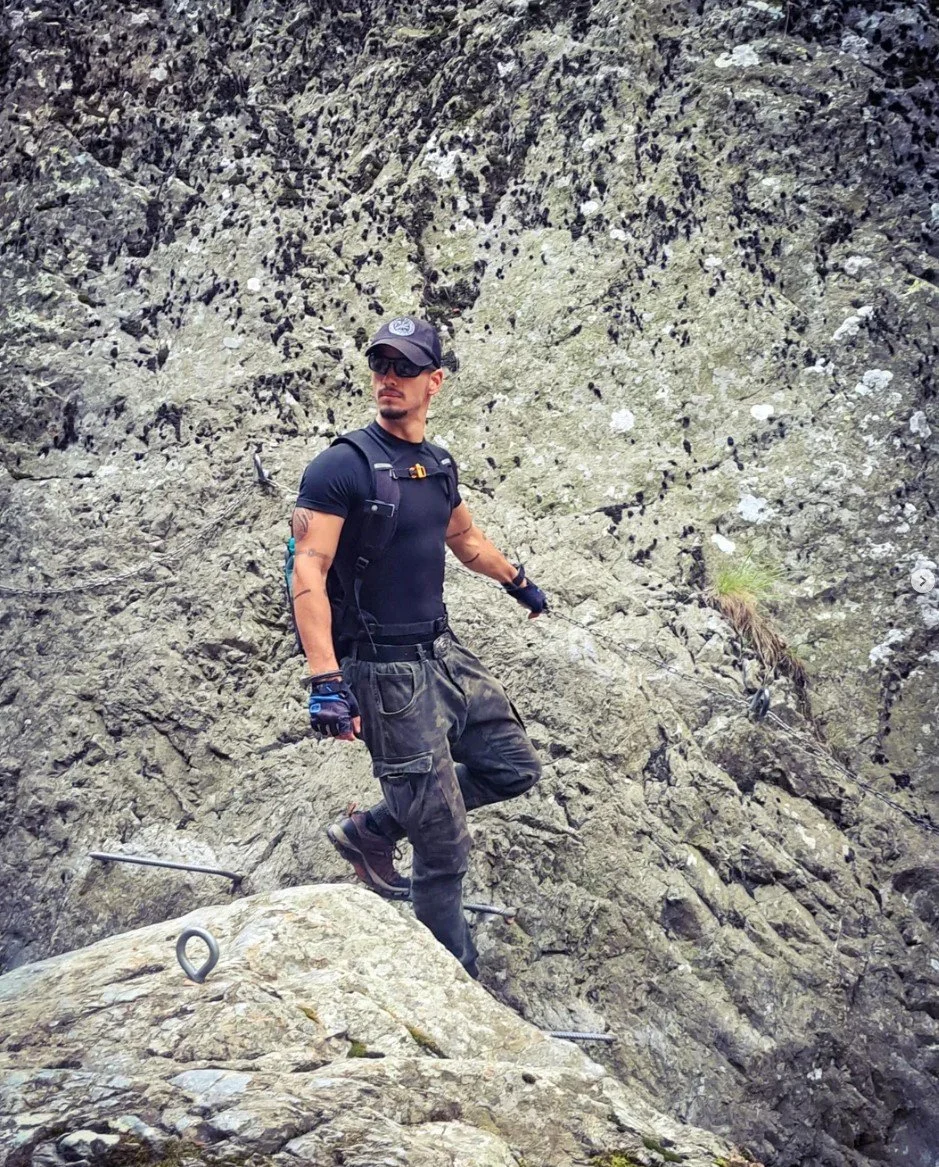 A man wearing sunglasses, a black shirt, camouflage pants, and a backpack, standing on a rocky mountain terrain.