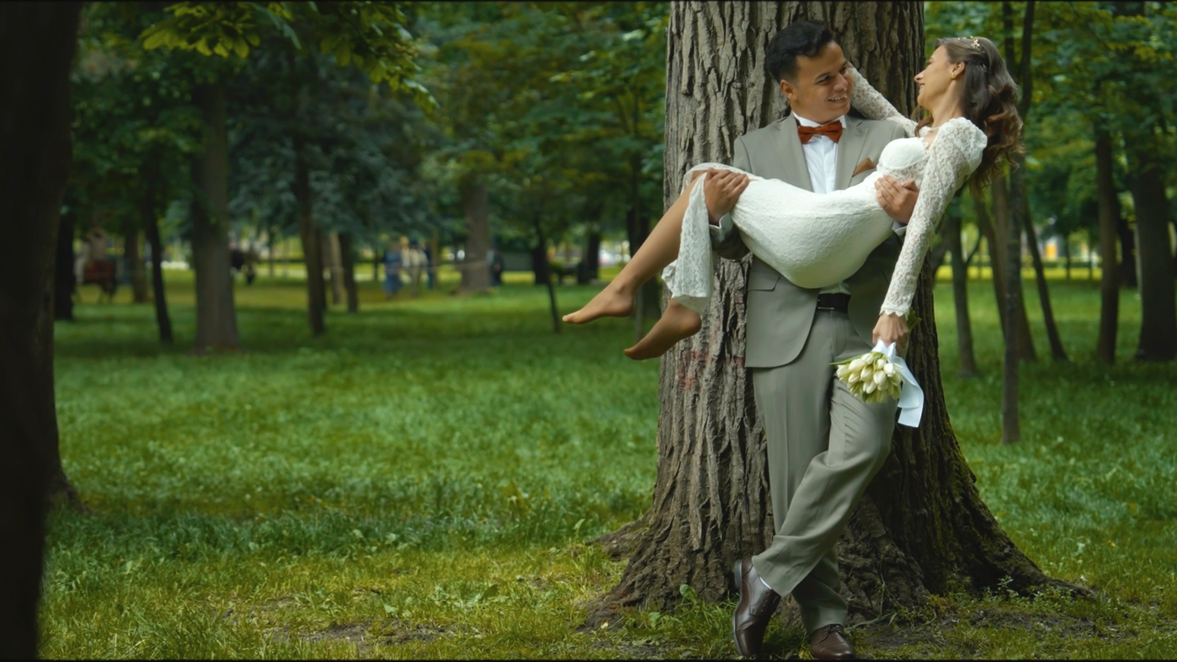 A man in a gray suit holding a woman in a white dress and lace, carrying her in a park with tall trees and green grass, smiling at each other. The woman holds a bouquet of white flowers.