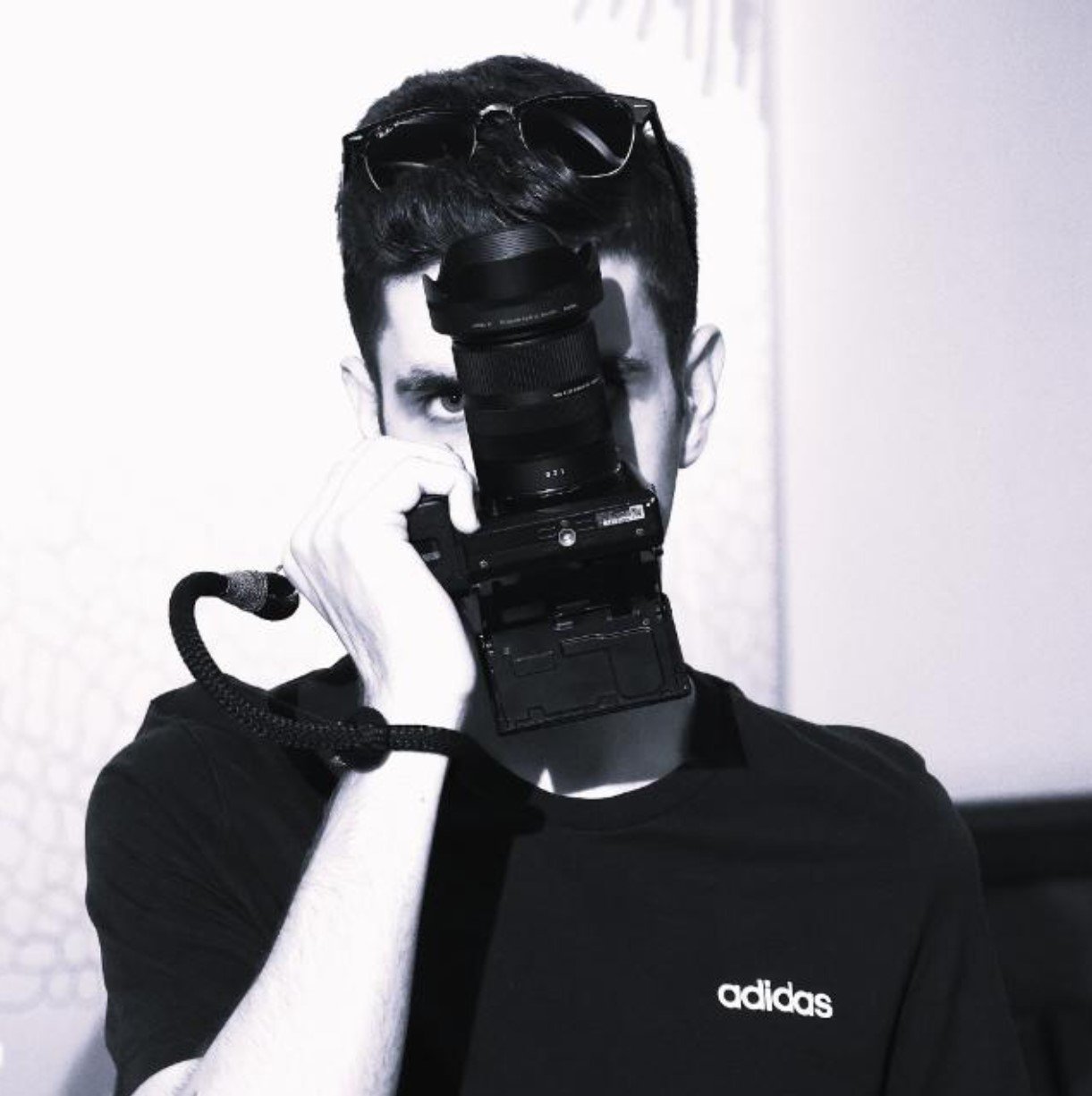 A person taking a photo with a camera, holding it up to their face, wearing a black Adidas shirt, with sunglasses resting on their head.