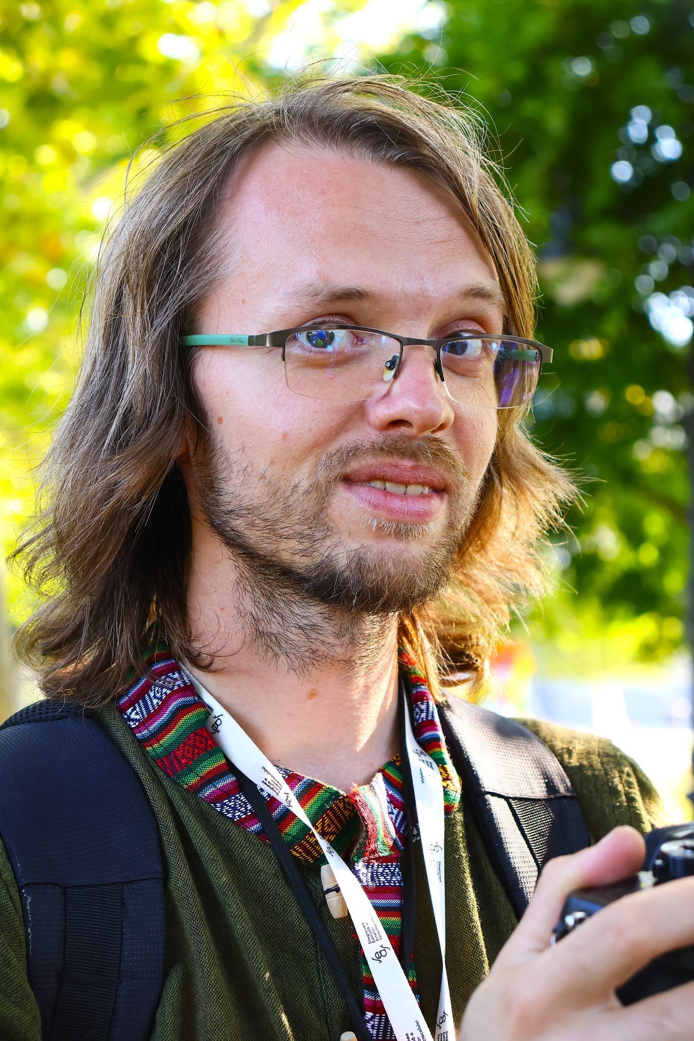 A man with shoulder-length brown hair, glasses, and a light beard and mustache holds a camera outdoors in front of green trees, wearing a colorful patterned shirt and a backpack.
