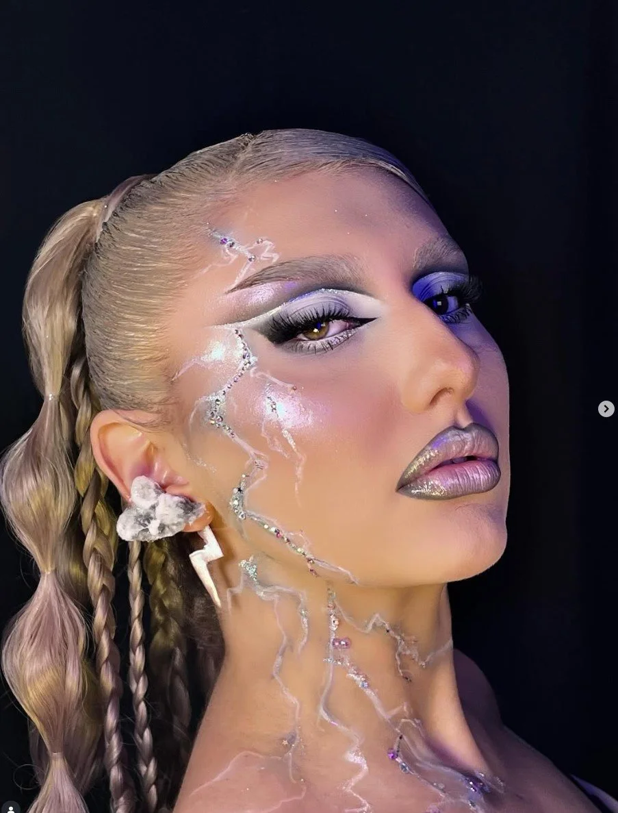 A woman with platinum blonde hair styled in braids, with metallic makeup, dramatic eye makeup, and shimmering details on her face and neck.
