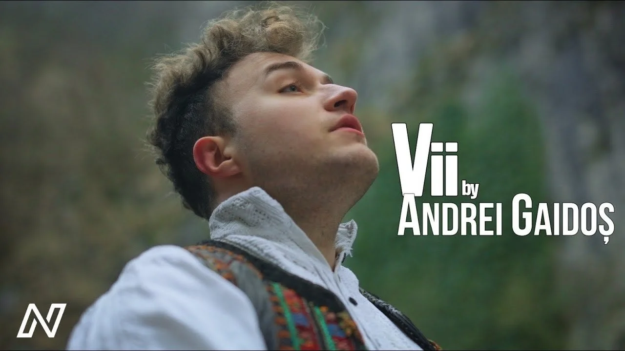 Close-up of a man with curly hair and a traditional embroidered shirt, outdoors in a forest, with text overlay reading "Vii by Andrei Gaidăș".