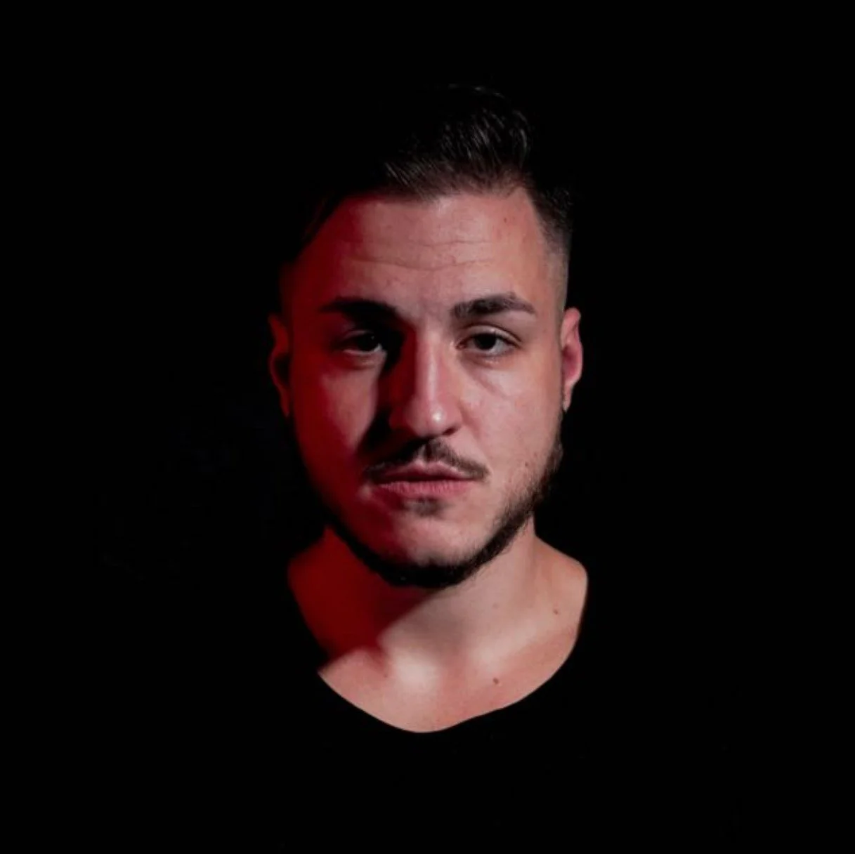 A young man with short dark hair, facial hair, and a serious expression. He is against a black background with red lighting highlighting the left side of his face.