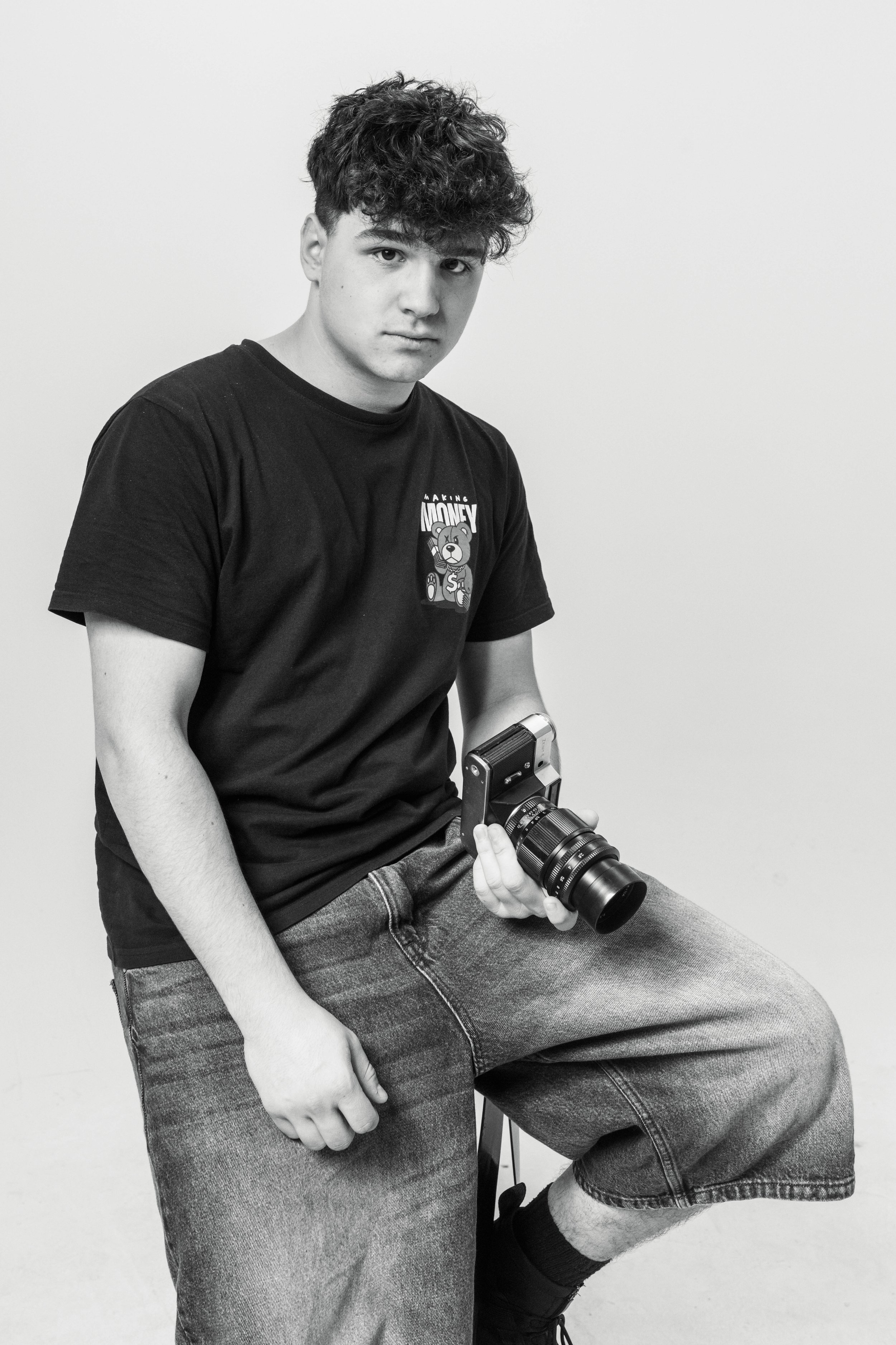 A young man with curly hair sitting on a stool, holding a vintage camera, wearing a black t-shirt and baggy jeans, against a plain background.