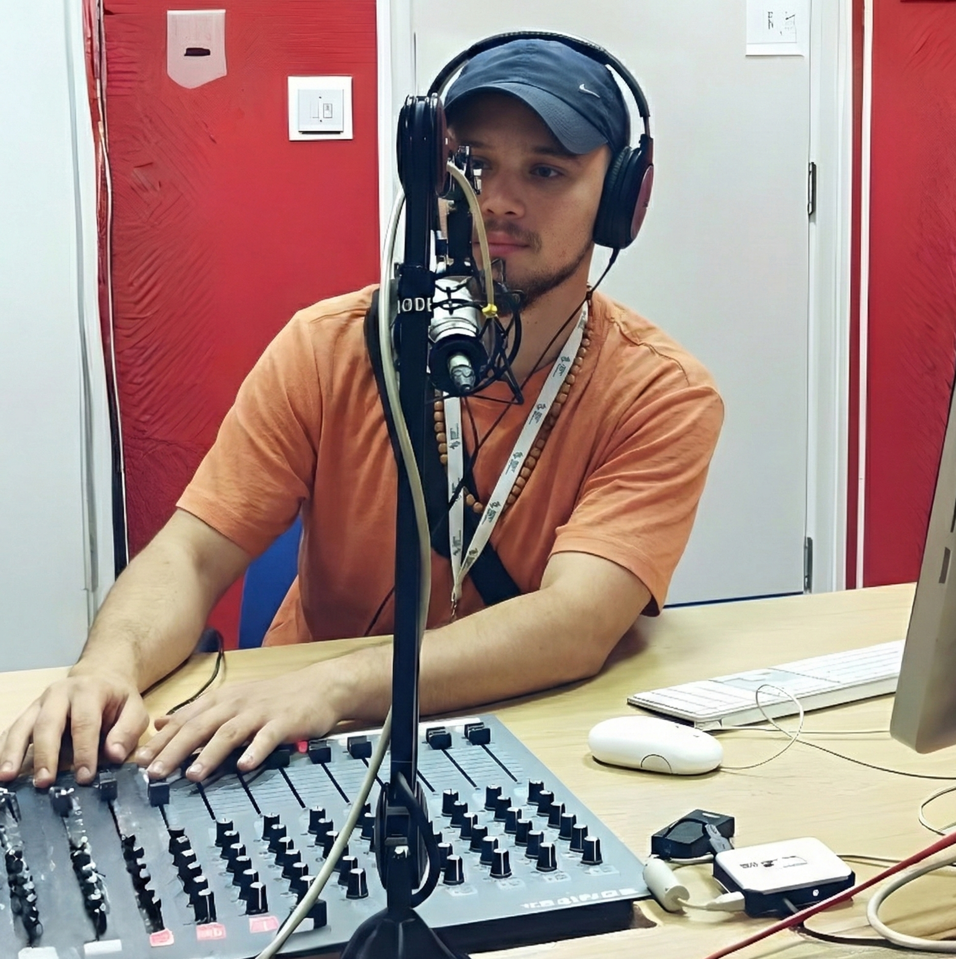 A young man with a beard, wearing a black cap, orange T-shirt, and headphones, is operating a recording microphone at a mixing console in a recording studio.