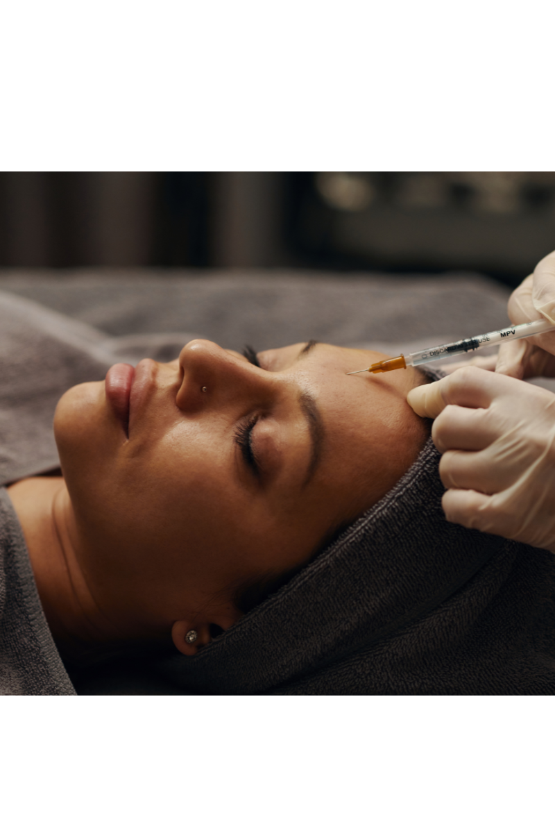 Frau liegt entspannt mit geschlossenen Augen, erhält eine kosmetische Behandlung mit einer Spritze im Gesicht, getragen von einer Person in Handschuhen.