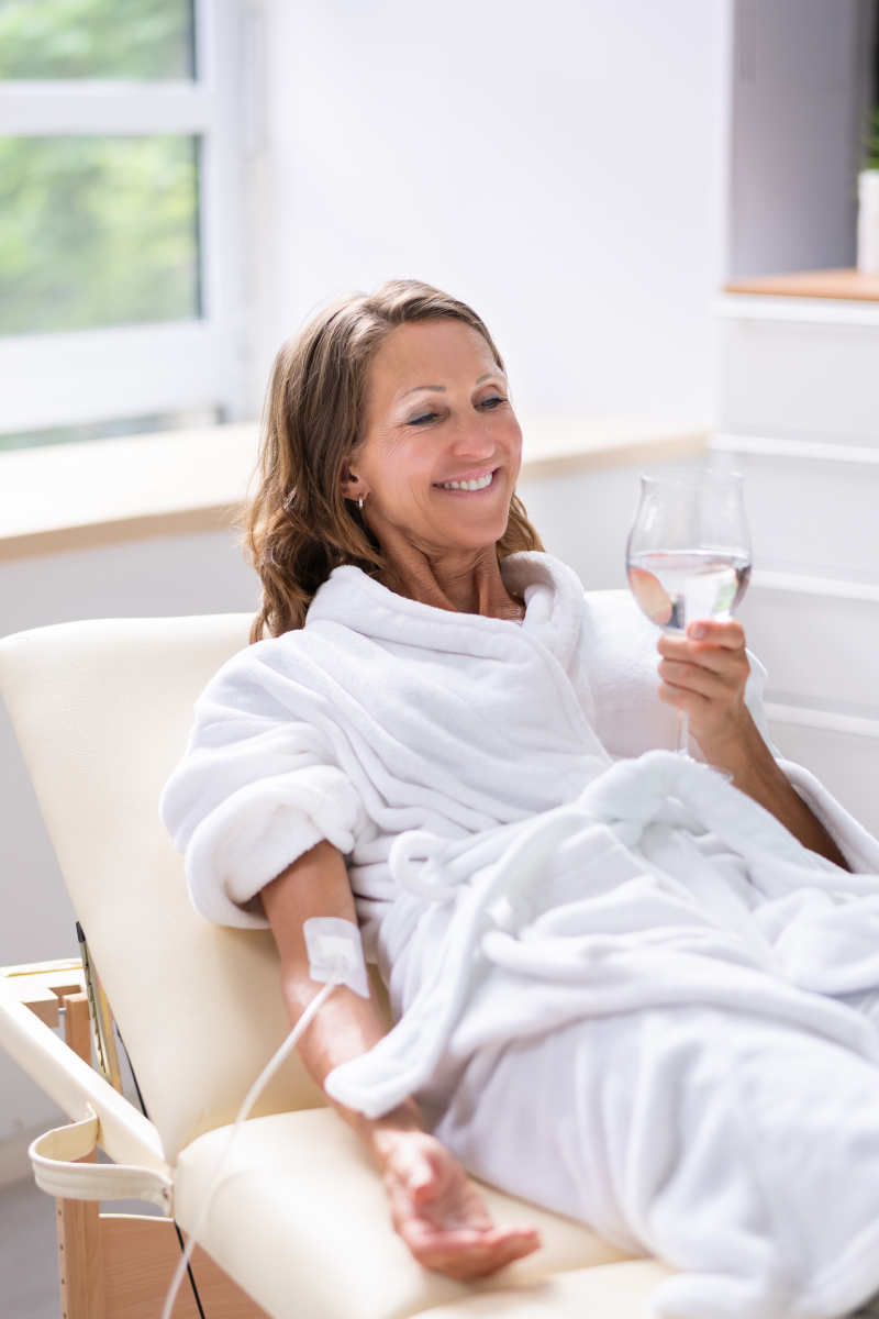 Eine lächelnde Frau in einem weißen Bademantel sitzt auf einem Krankenhausbett und hält ein Glas Wasser.