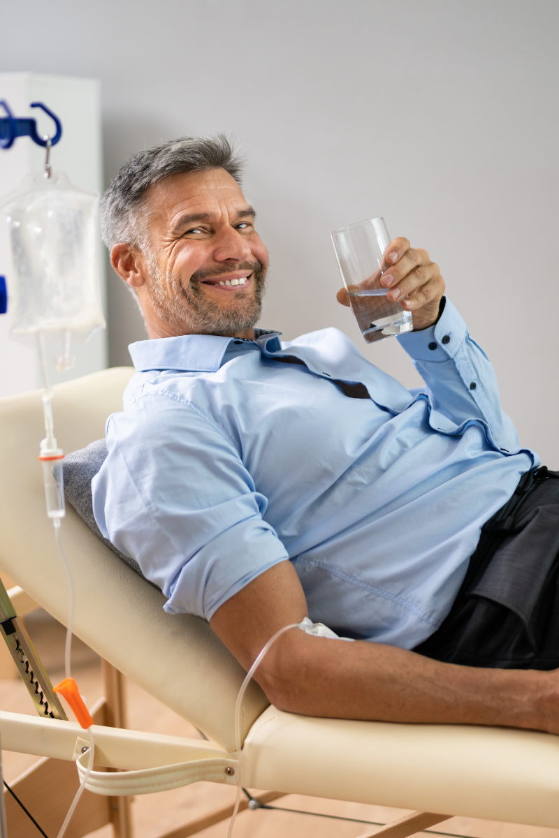 Ein Mann liegt auf einer Liege im Krankenhaus, hält ein Glas Wasser und lächelt in die Kamera, während eine Infusionsflasche im Hintergrund hängt.