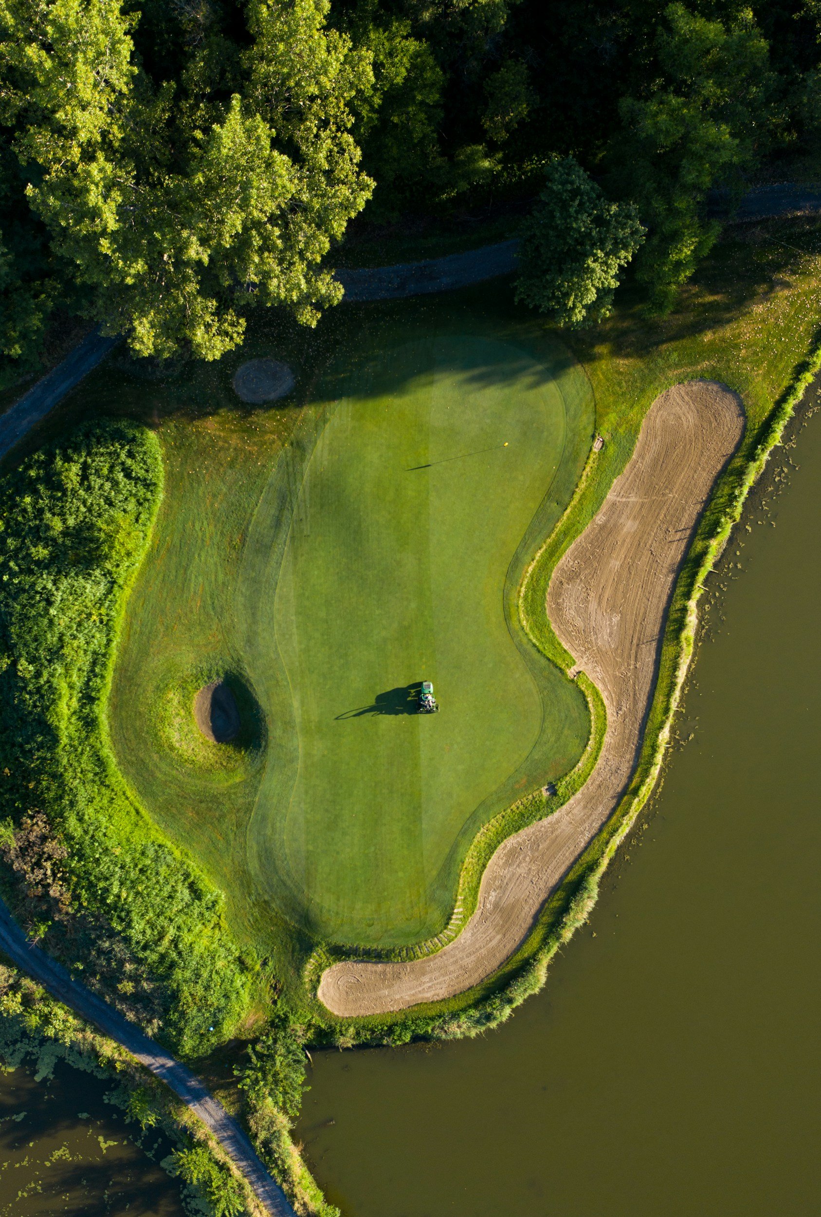 Aerial view of a golf course green surrounded by trees, water, and sand bunkers, with a person on a golf cart on the green.