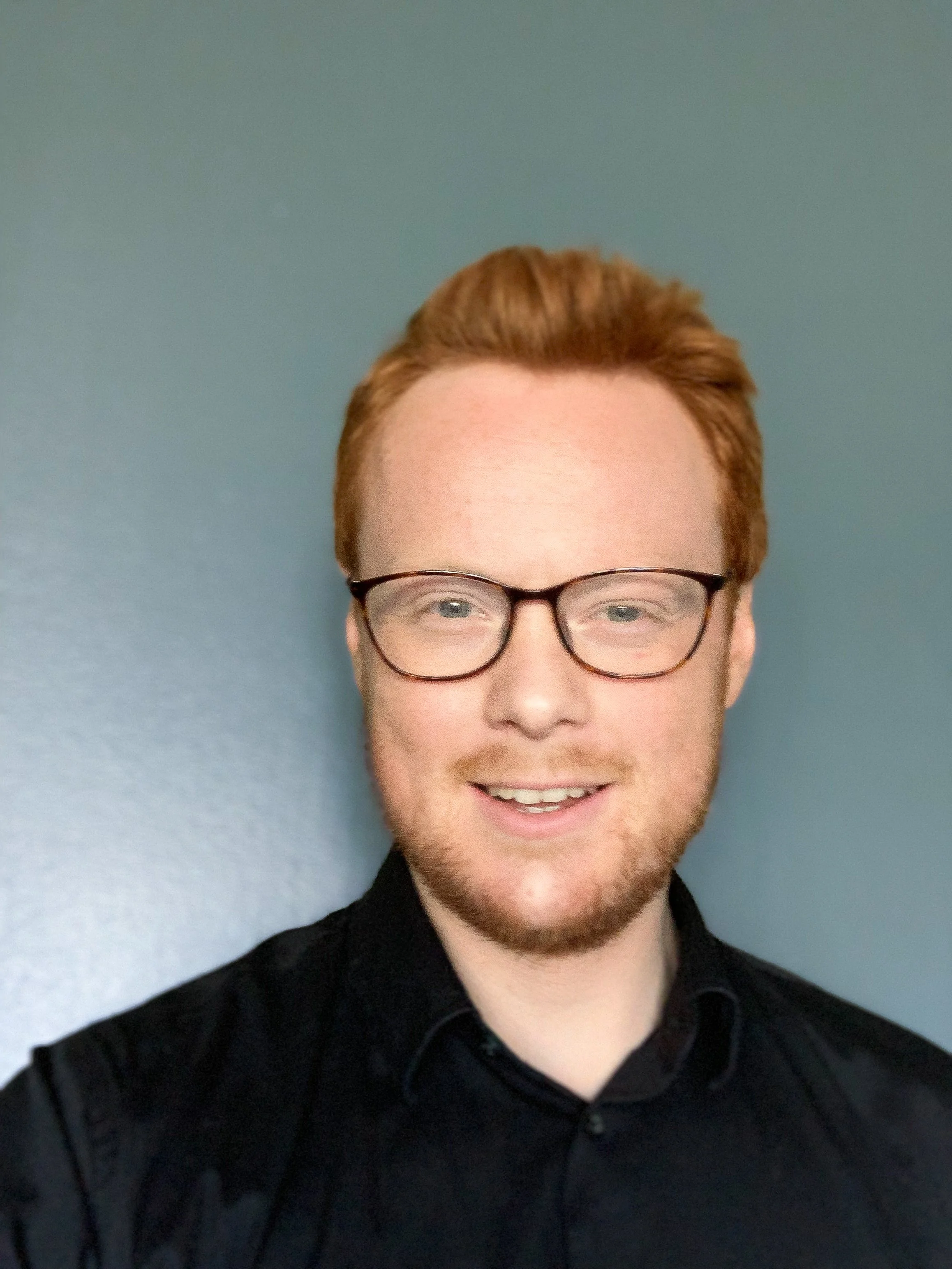 A young man with red hair, glasses, and a beard smiling against a blue wall.