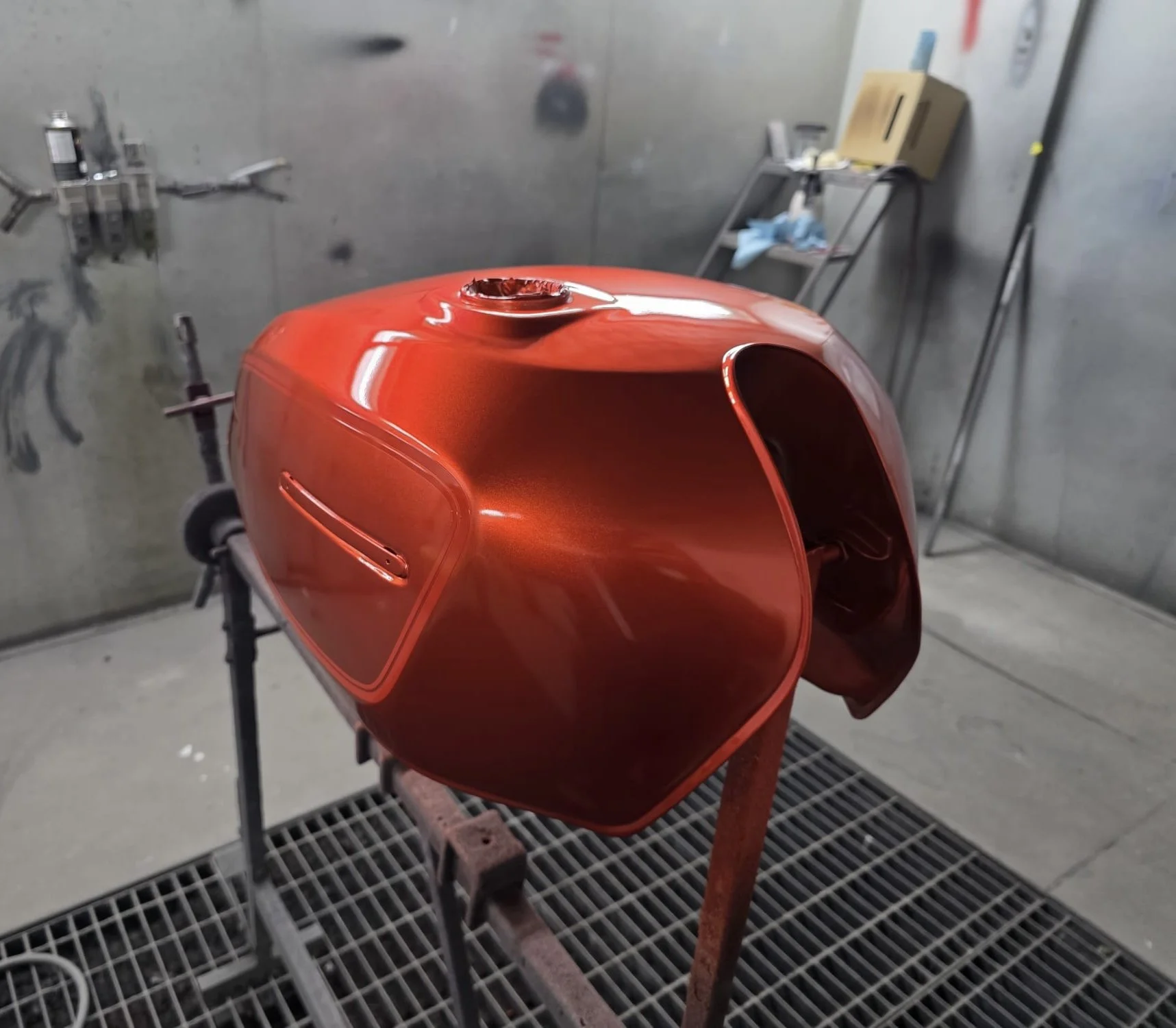 A red motorcycle fuel tank in a paint booth with a metallic finish, mounted on a stand, with tools and equipment in the background.