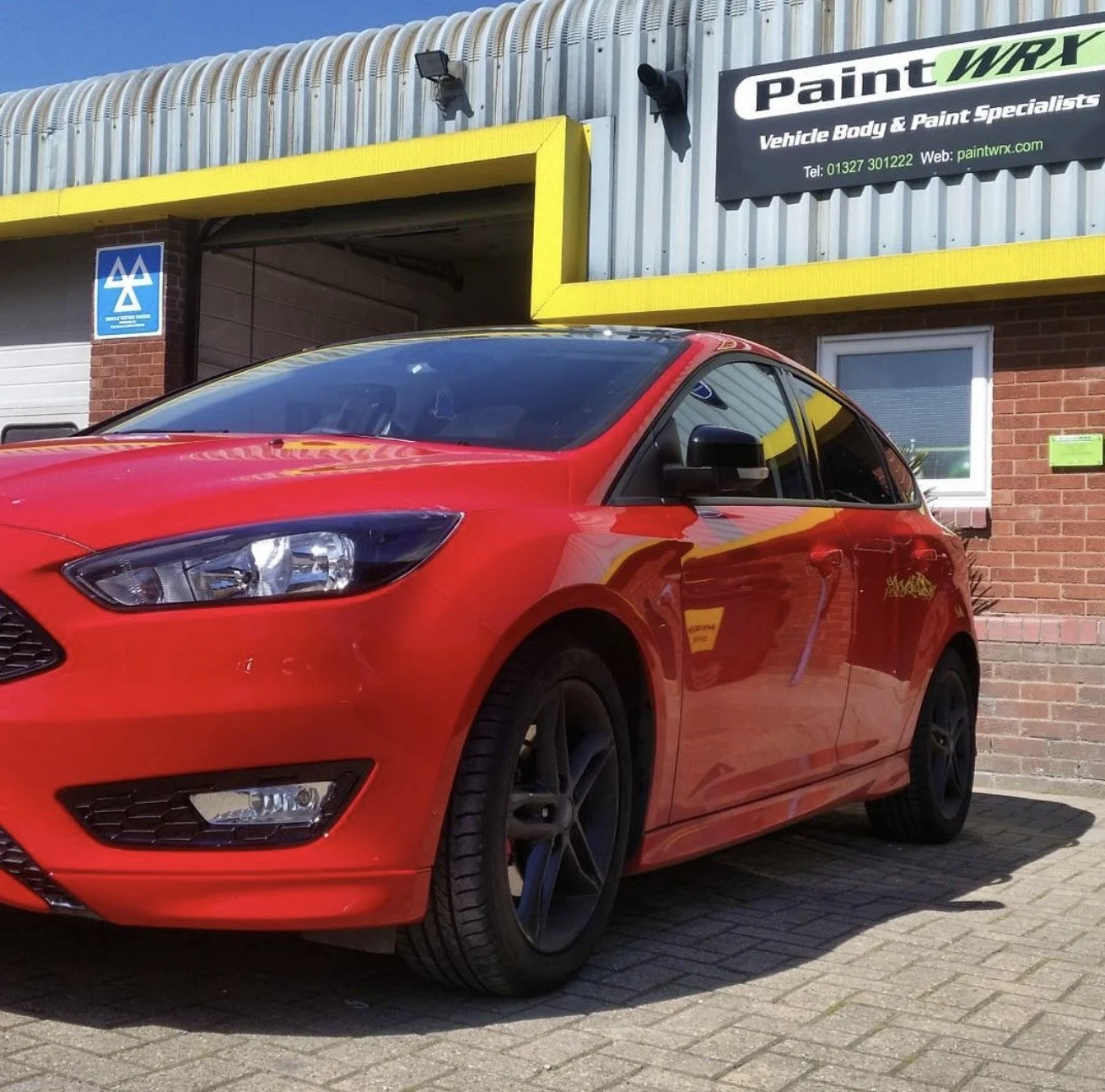 Red hatchback car parked outside a building with signs and brick wall, sunny day.