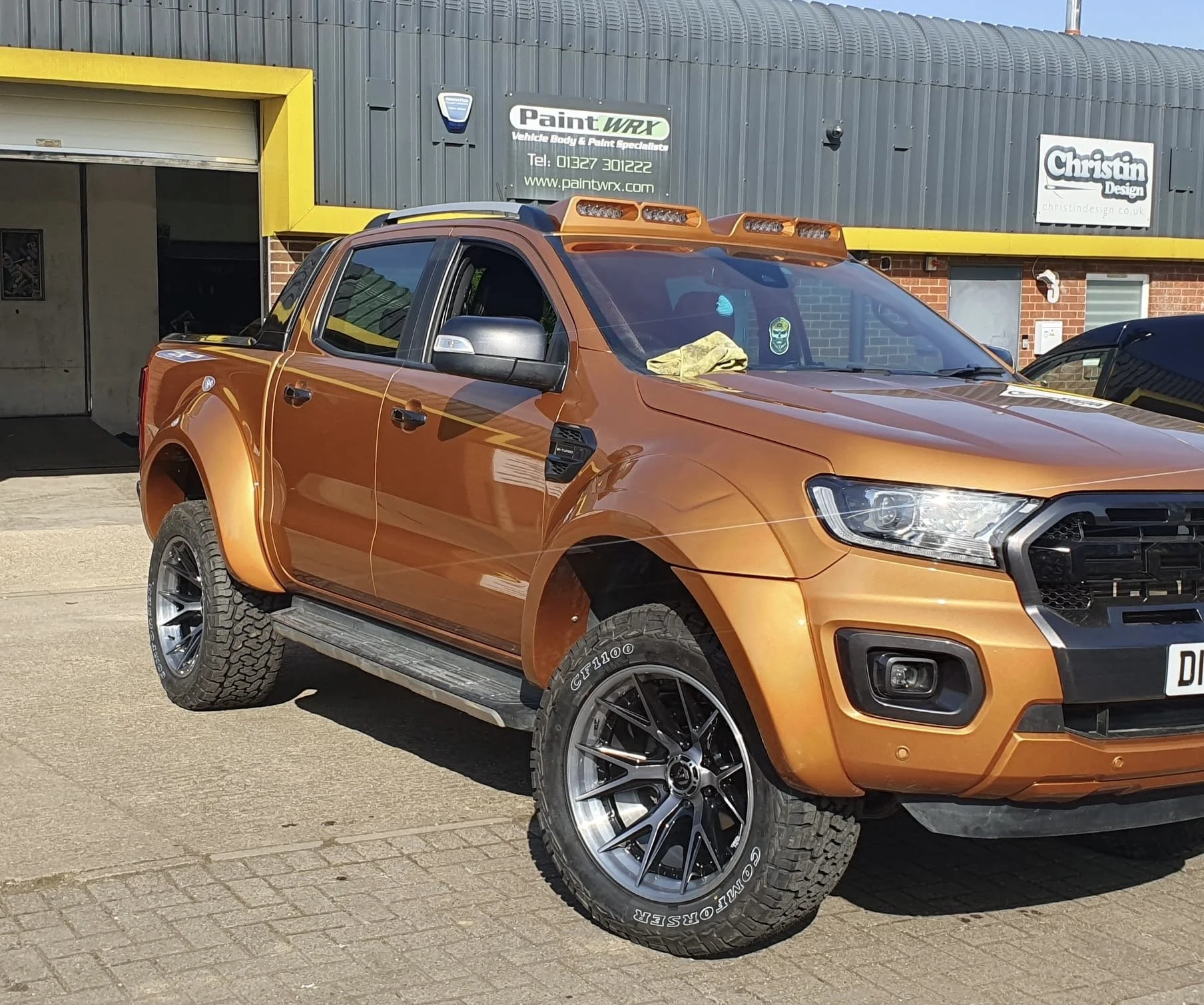 An orange pickup truck with black wheels parked outside a building with yellow and gray accents, some signs on the building, and other vehicles nearby.