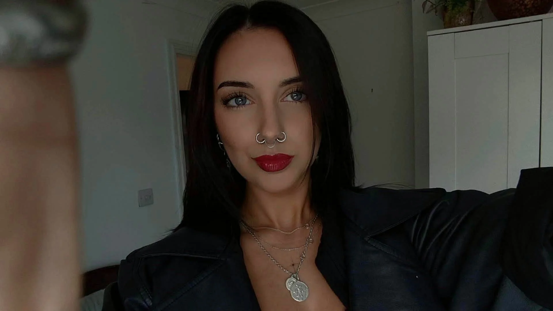 A woman with dark hair, blue eyes, red lipstick, and tattoos, taking a selfie indoors in front of a white cabinet, wearing layered silver necklaces and a black leather jacket.