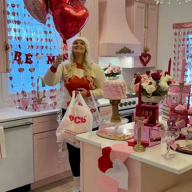 A woman with long blonde hair holding a heart-shaped balloon and a bag, celebrating Valentine's Day in a pink-themed decorated kitchen with flowers, heart-shaped cutouts, and pink cakes.