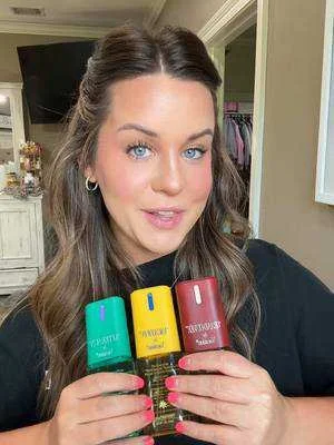 A woman with long brown hair, blue eyes, and hoop earrings holds three bottles of nail polish in green, yellow, and red colors.