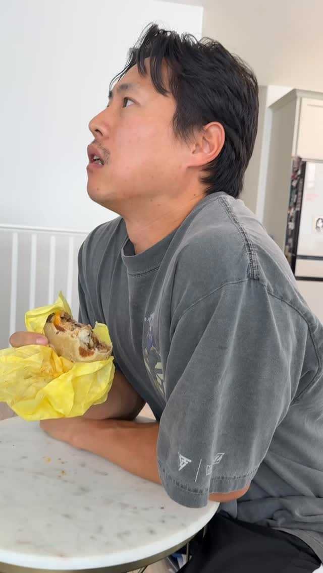 A man with short black hair and wearing a gray T-shirt sitting at a white table, holding a partially eaten hot dog wrapped in yellow paper, with a surprised or confused expression on his face.