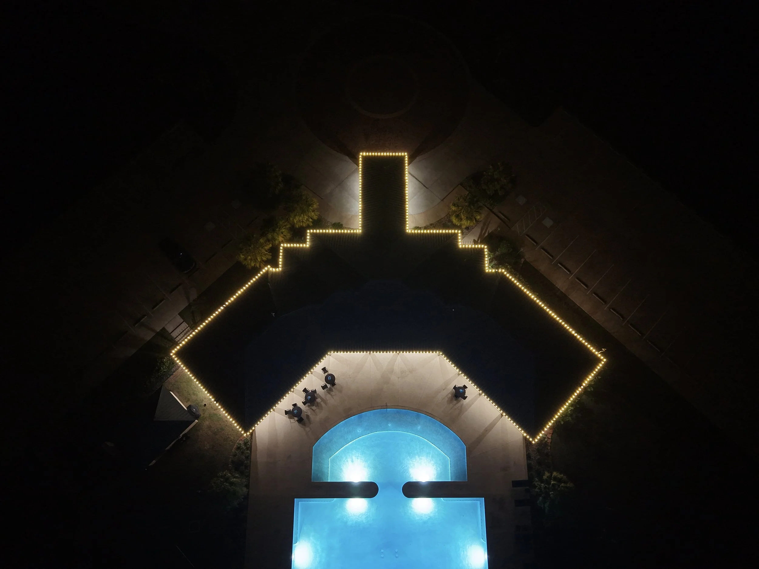 Aerial nighttime view of a swimming pool shaped like the United States outline, illuminated by yellow lights around the perimeter, with a smaller, circular blue-lit pool at the bottom. There are a few chairs and trees outside.