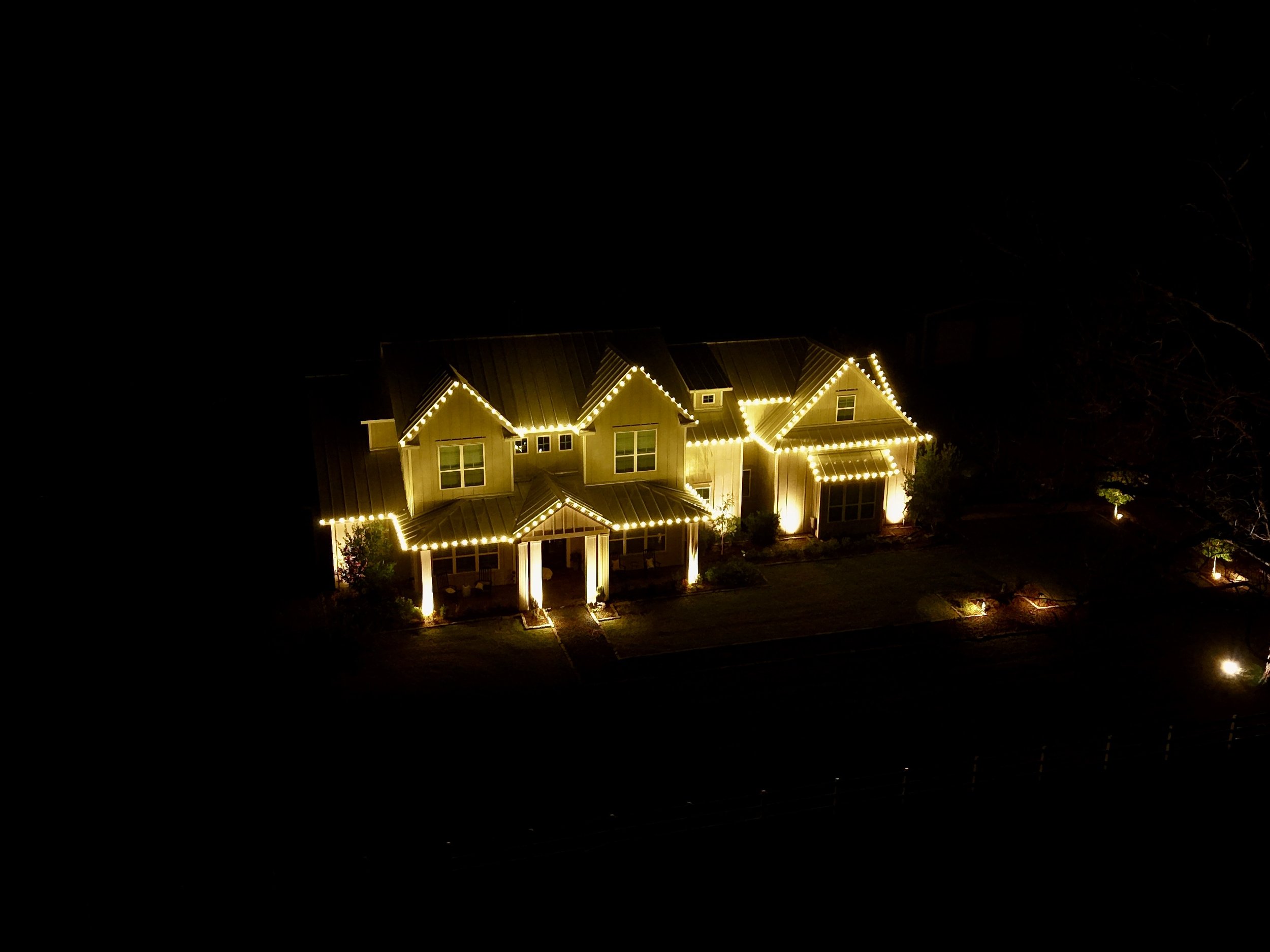 Night view of a house decorated with yellow string lights outlining the roof and windows, with landscape lighting illuminating the yard.
