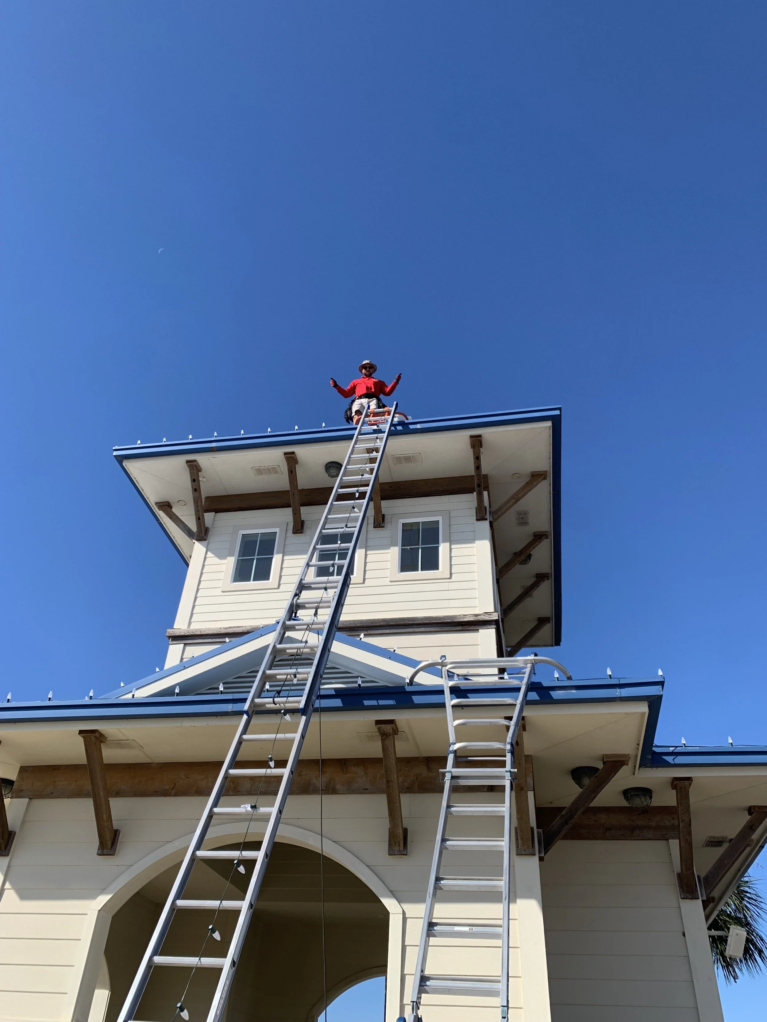 Person standing on a rooftop of a house, holding a ladder, against a clear blue sky.