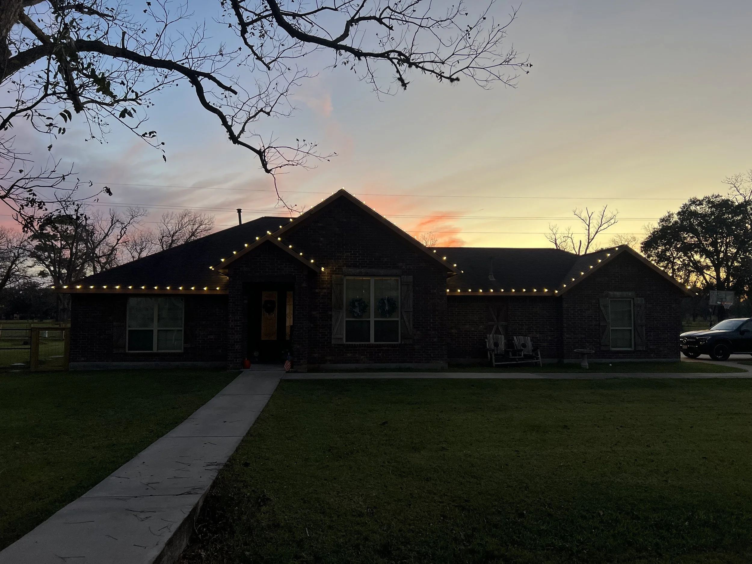 A brick house decorated with string lights along the roofline during sunset, with a front walkway, lawn, and a few trees and a vehicle in the background.