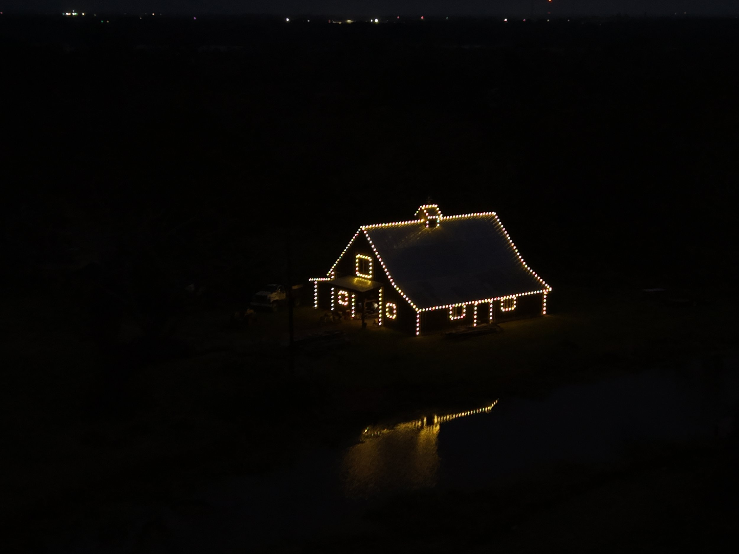 A house decorated with Christmas lights, reflecting on a nearby body of water, at night.