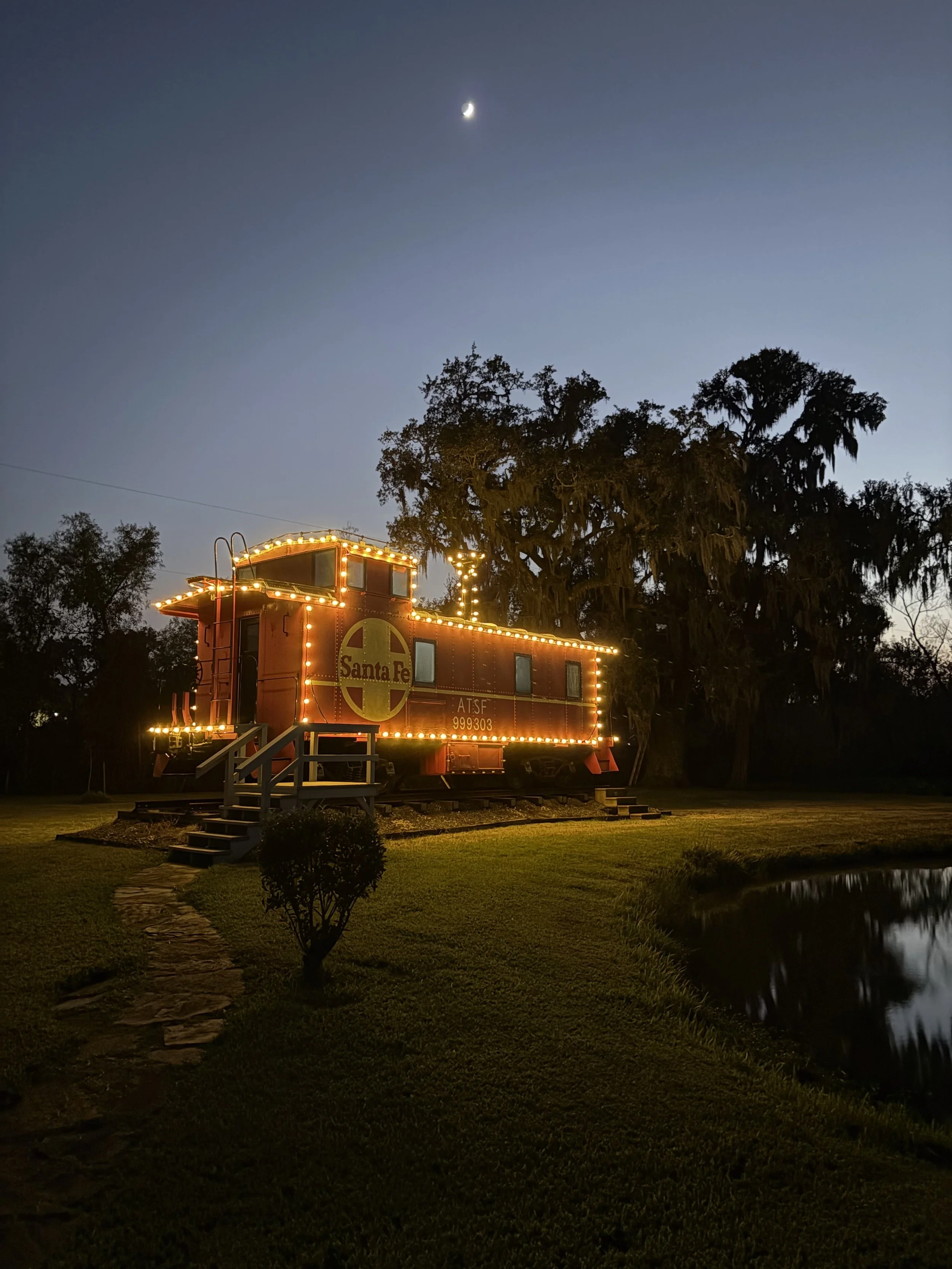Nighttime scene with a small vintage train decorated with yellow string lights, labeled "Santa Fe," on a grassy area with trees and a small pond nearby, under a clear sky with a crescent moon visible.