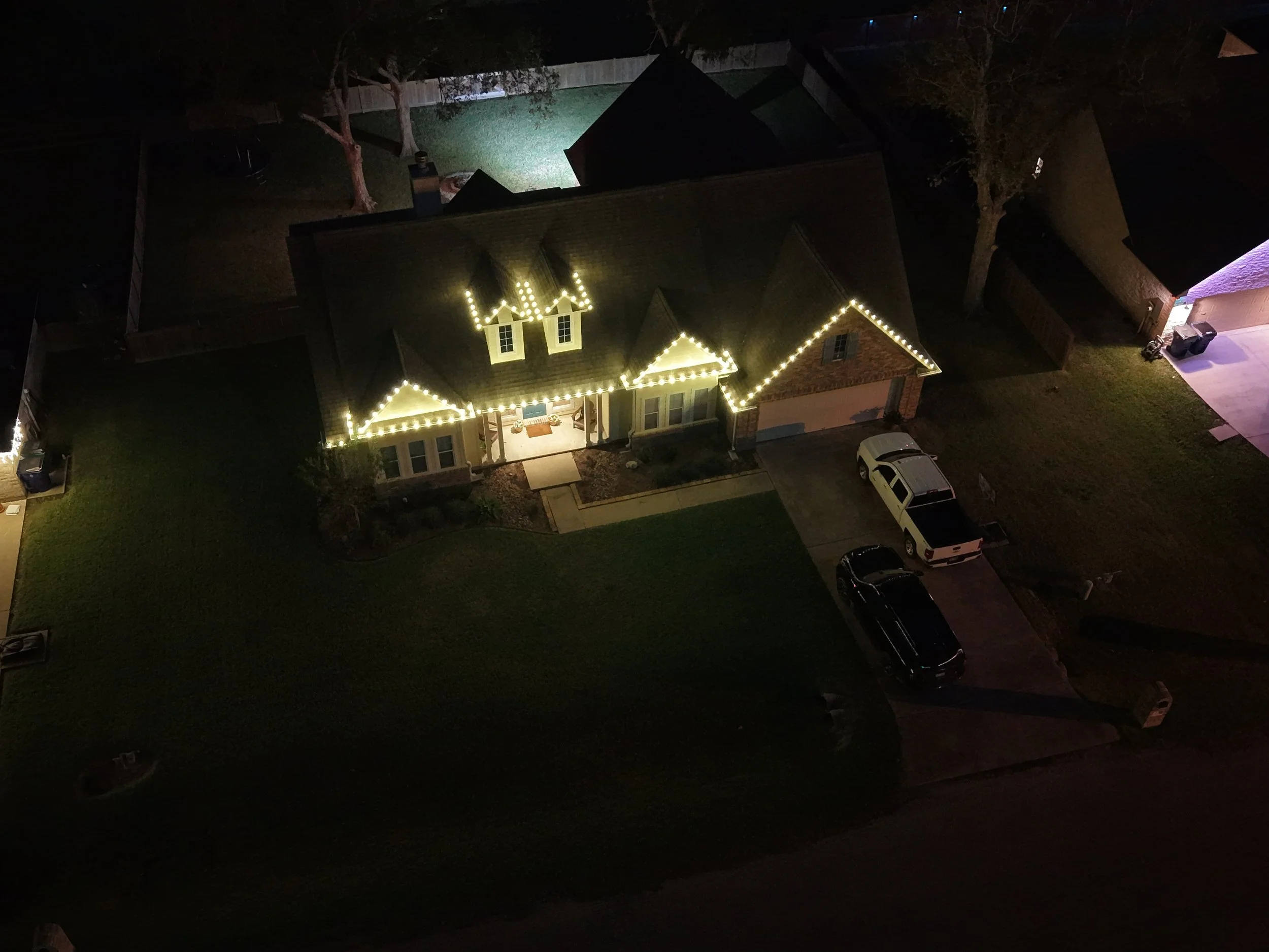 Aerial nighttime view of a house decorated with string lights on the roof and porch, with three parked cars in the driveway and a grassy yard.