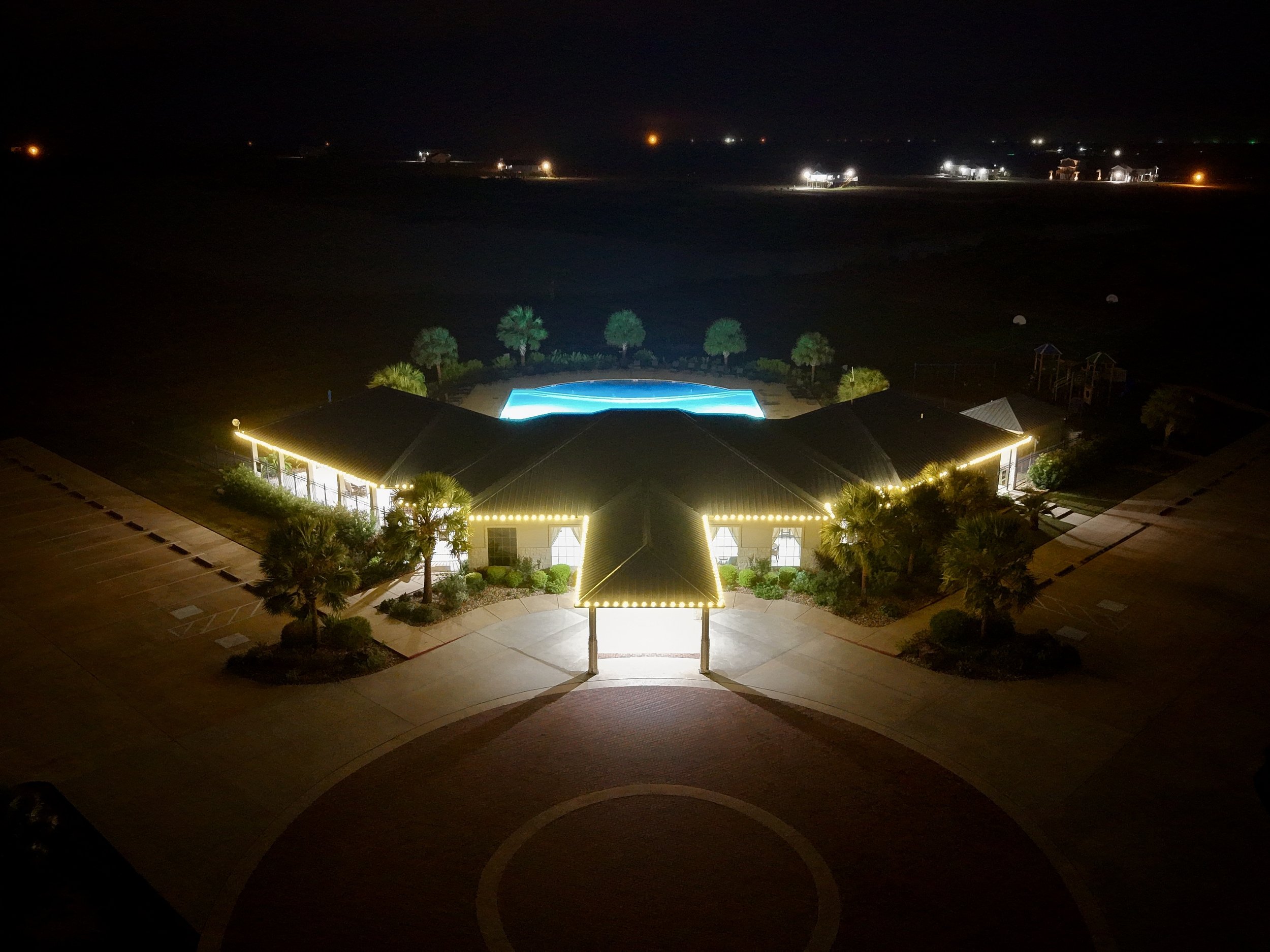 Nighttime aerial view of a building with string lights along the roof, surrounded by palm trees, with a lit swimming pool behind it and an empty parking lot in the foreground.