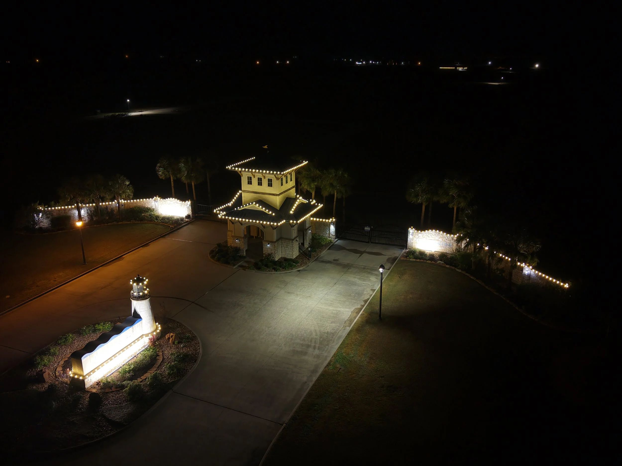 Nighttime view of a well-lit entrance to a property, featuring a small lighthouse sign and a gated gate with additional lighting along the fence, surrounded by trees and open land in the background.