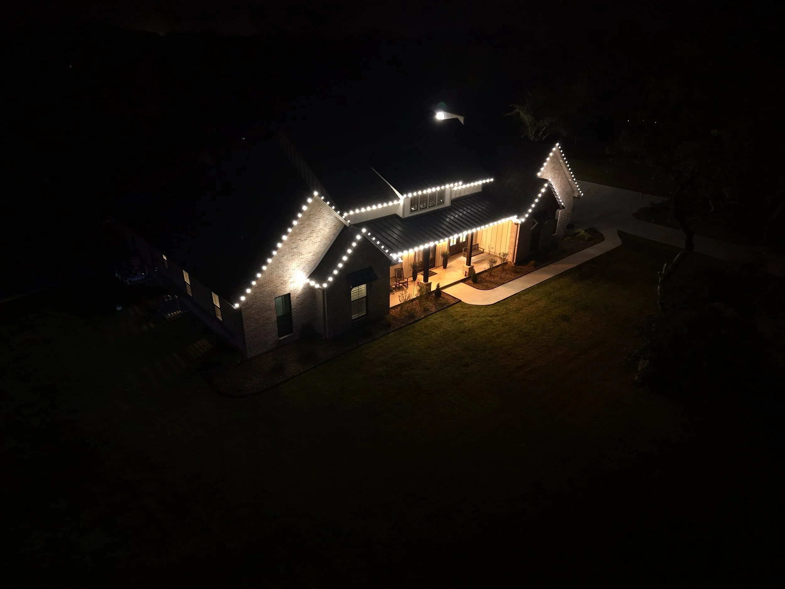 A house decorated with string lights outlining the roof, at night. The house has a front porch with potted plants and a walkway leading to it. The yard surrounds the house, with trees and a driveway visible.