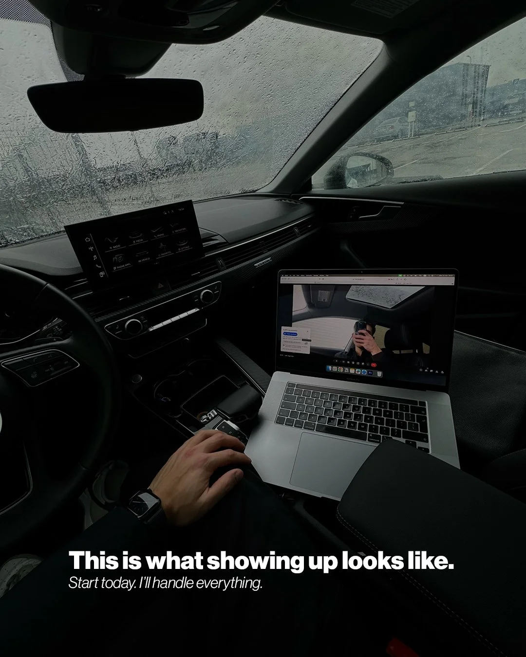 Inside a car during rainy weather, with a laptop on the passenger seat showing a person taking a selfie, and the driver's hand resting on a device. The windshield shows rain and a parking lot outside.