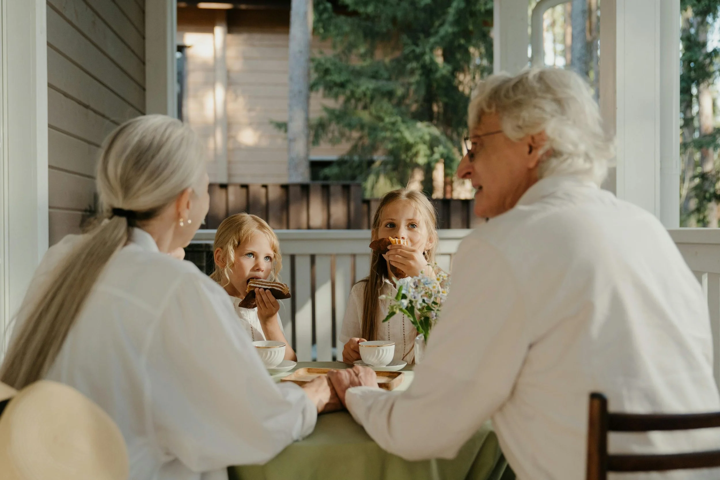Grandparents and two young girls sitting at a dining table enjoying breakfast or tea, with two cups and a small flower arrangement, in a bright room with large windows showing trees outside.