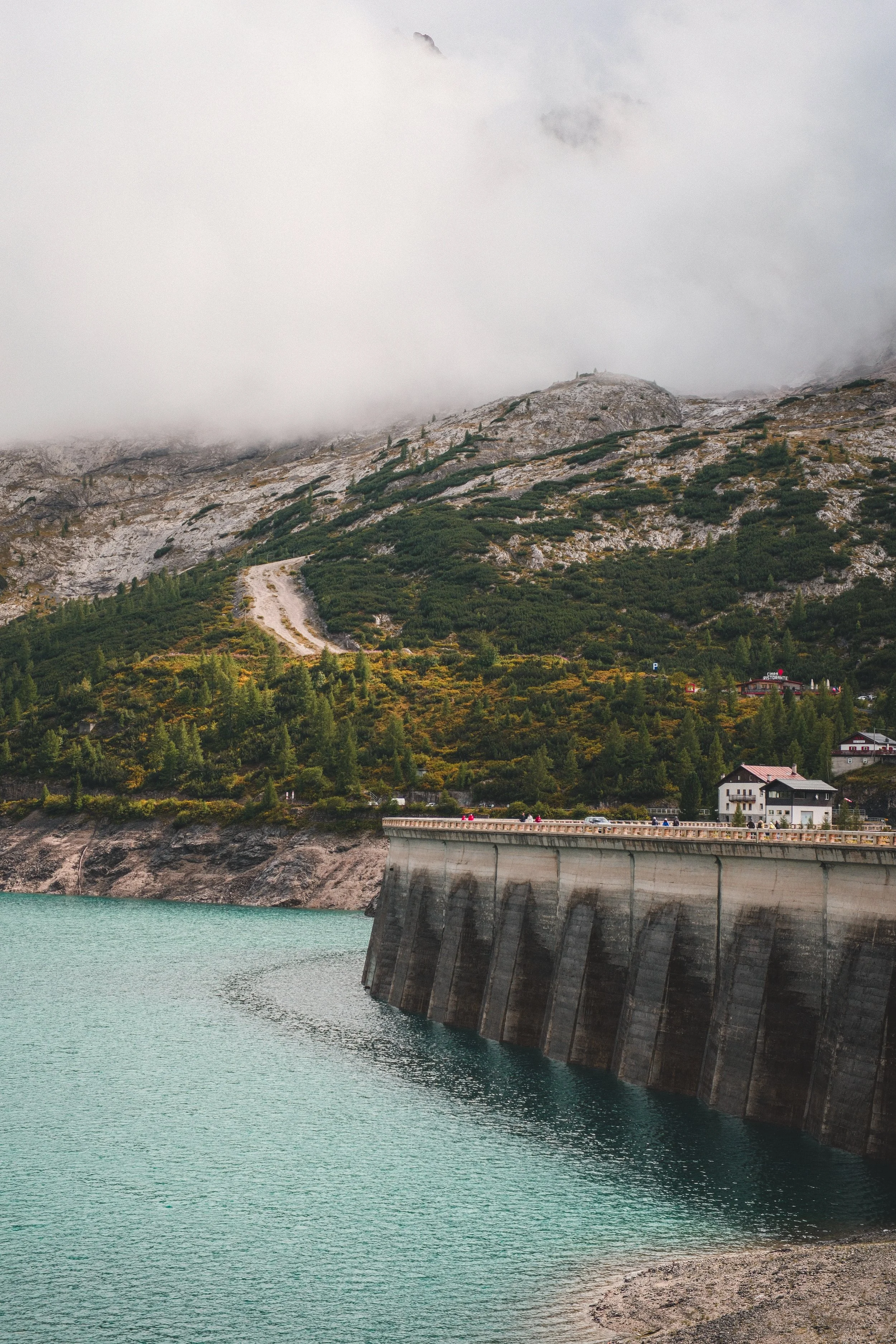 A dam next to a body of water with mountains in the background, partly covered with low-hanging clouds.