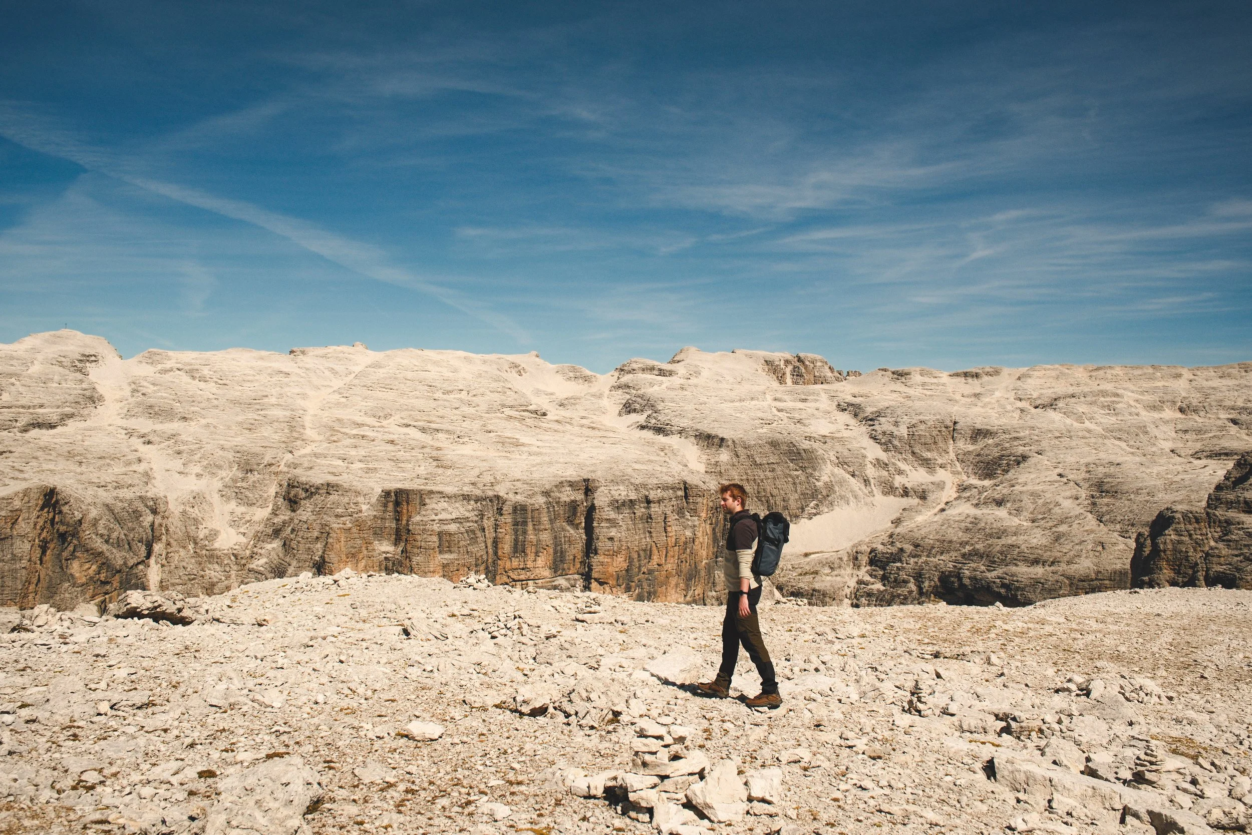 A hiker with a large backpack walking in a rocky, mountainous landscape under a blue sky with wispy clouds.