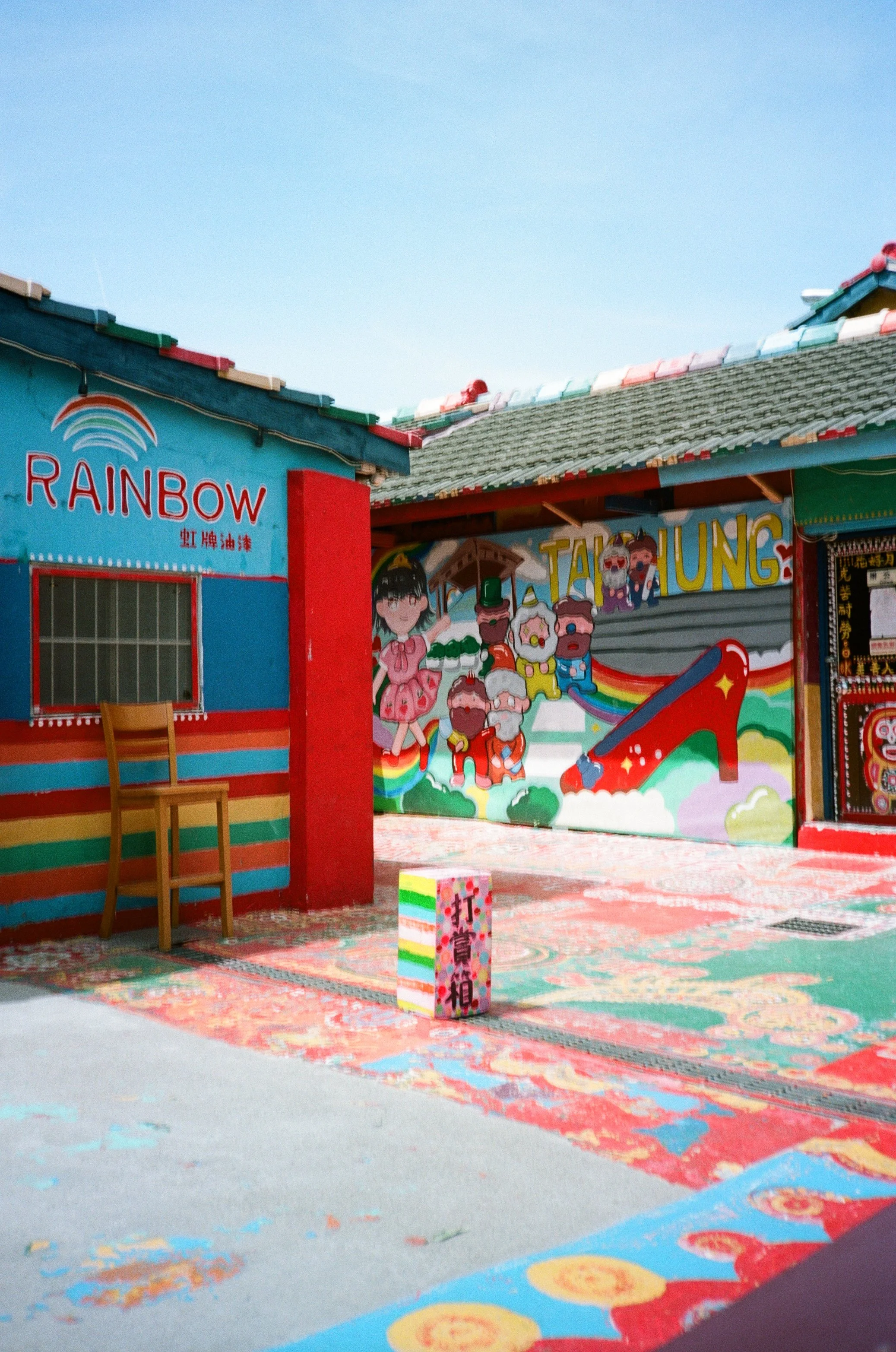 Colorful mural with cartoon characters, rainbow, and the word 'Tatung', part of a vibrant, festive setting with paint splatters on the ground.