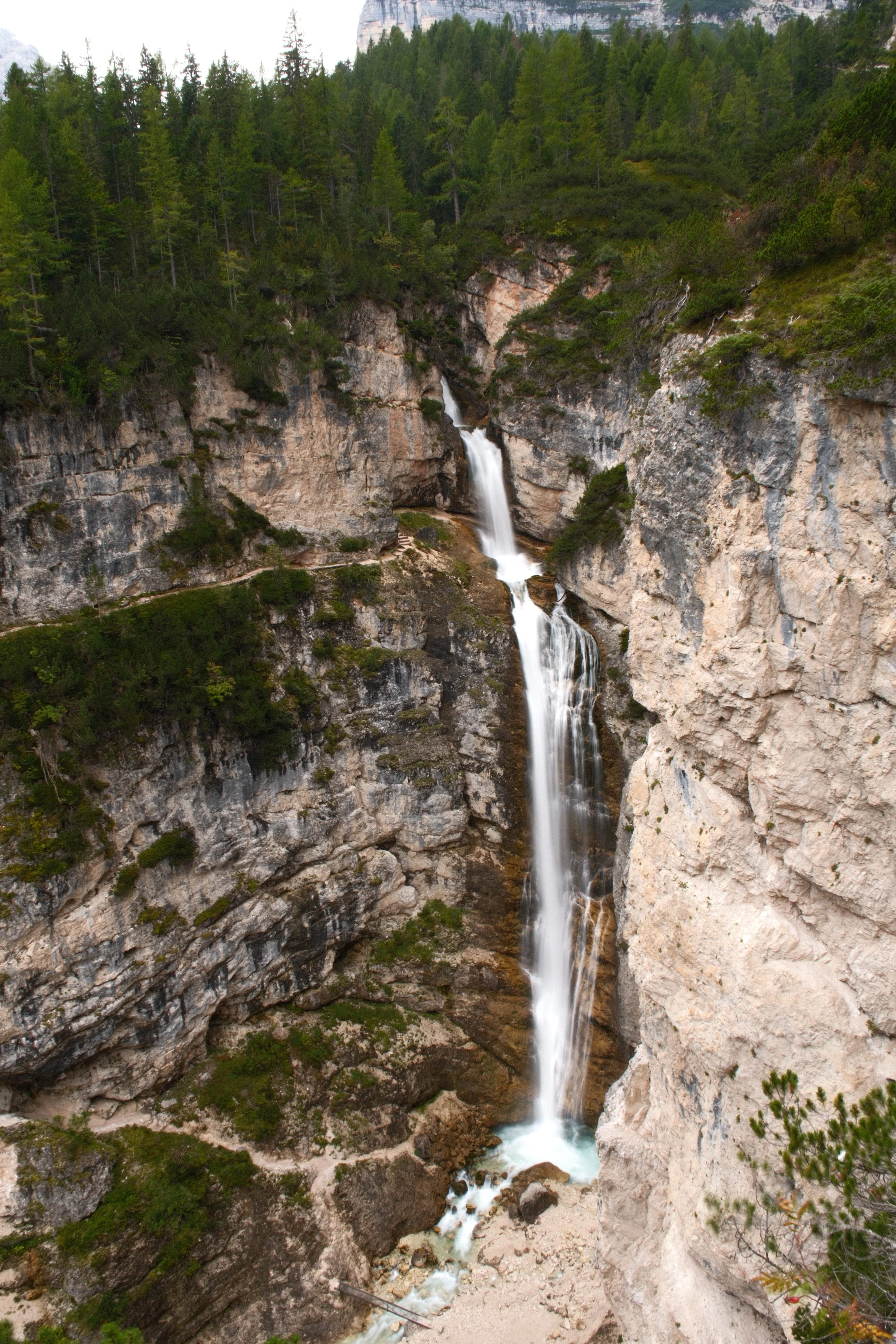 A tall waterfall cascades down a steep rocky cliff surrounded by dense forest of green coniferous trees in a mountainous area.