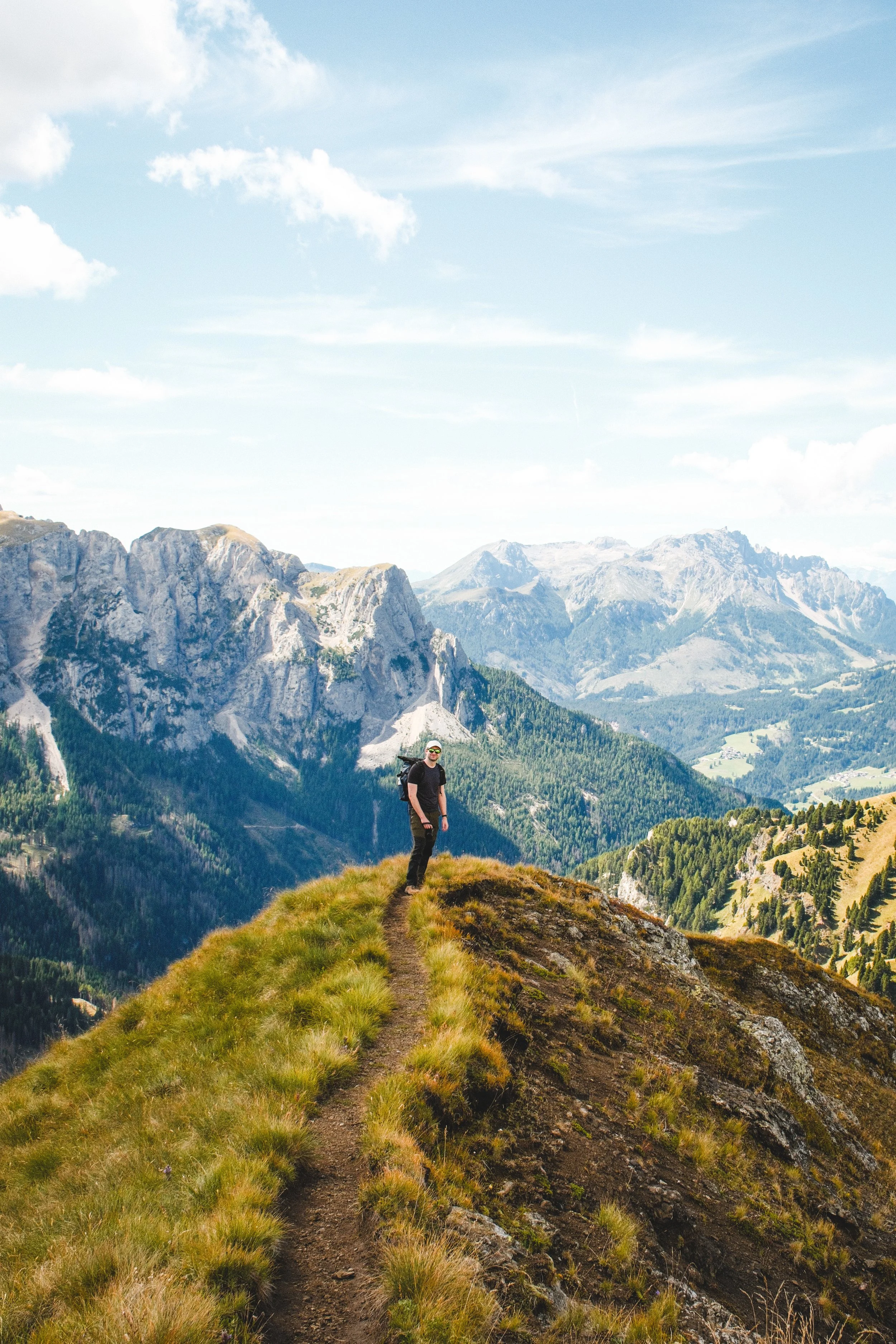 A hiker walking along a mountain trail surrounded by green grass, with rugged mountain peaks in the background under a partly cloudy sky.