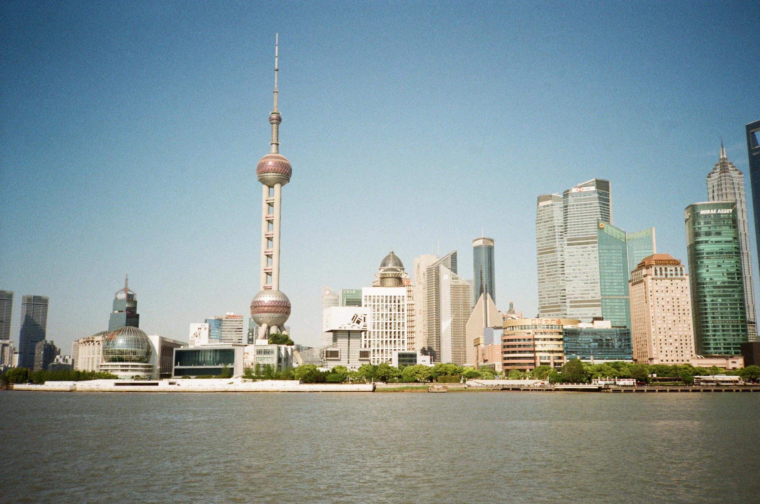 City skyline featuring the Oriental Pearl Tower with its distinctive spheres, surrounded by modern skyscrapers, seen from across a river on a clear day.