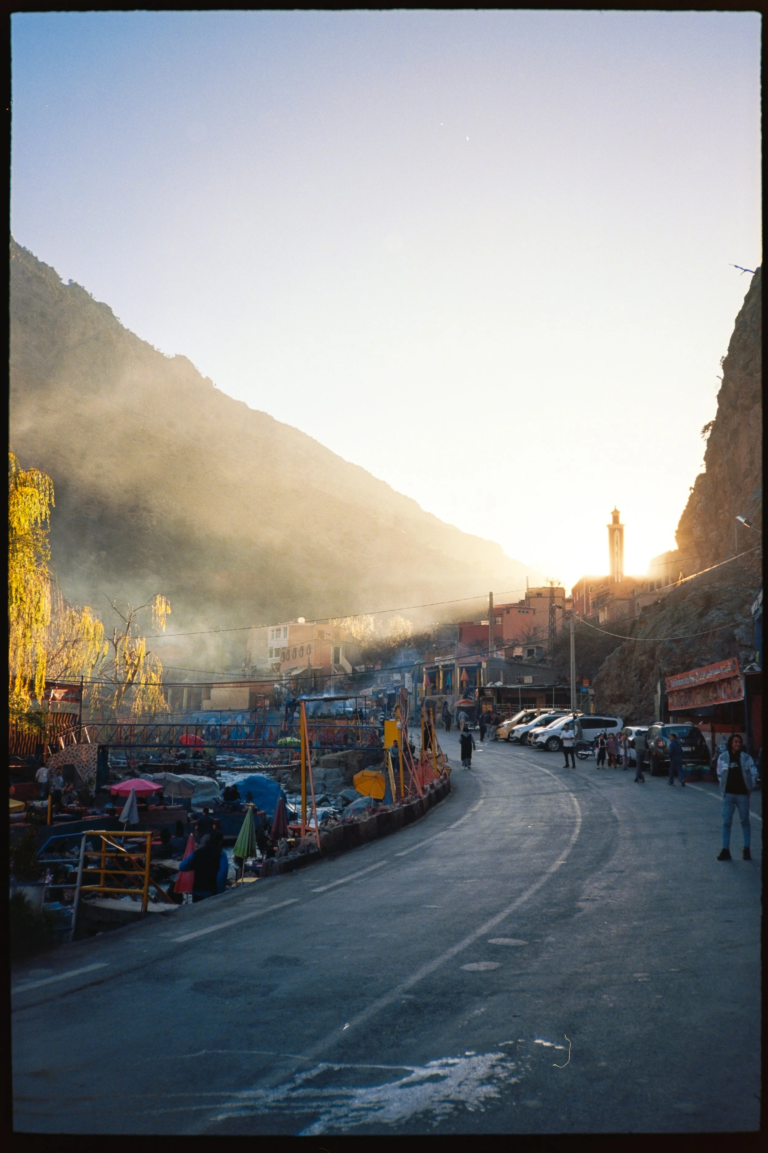A sunlit street in a small town at the base of a mountain, with people walking, parked cars, and a marketplace with umbrellas and tents.