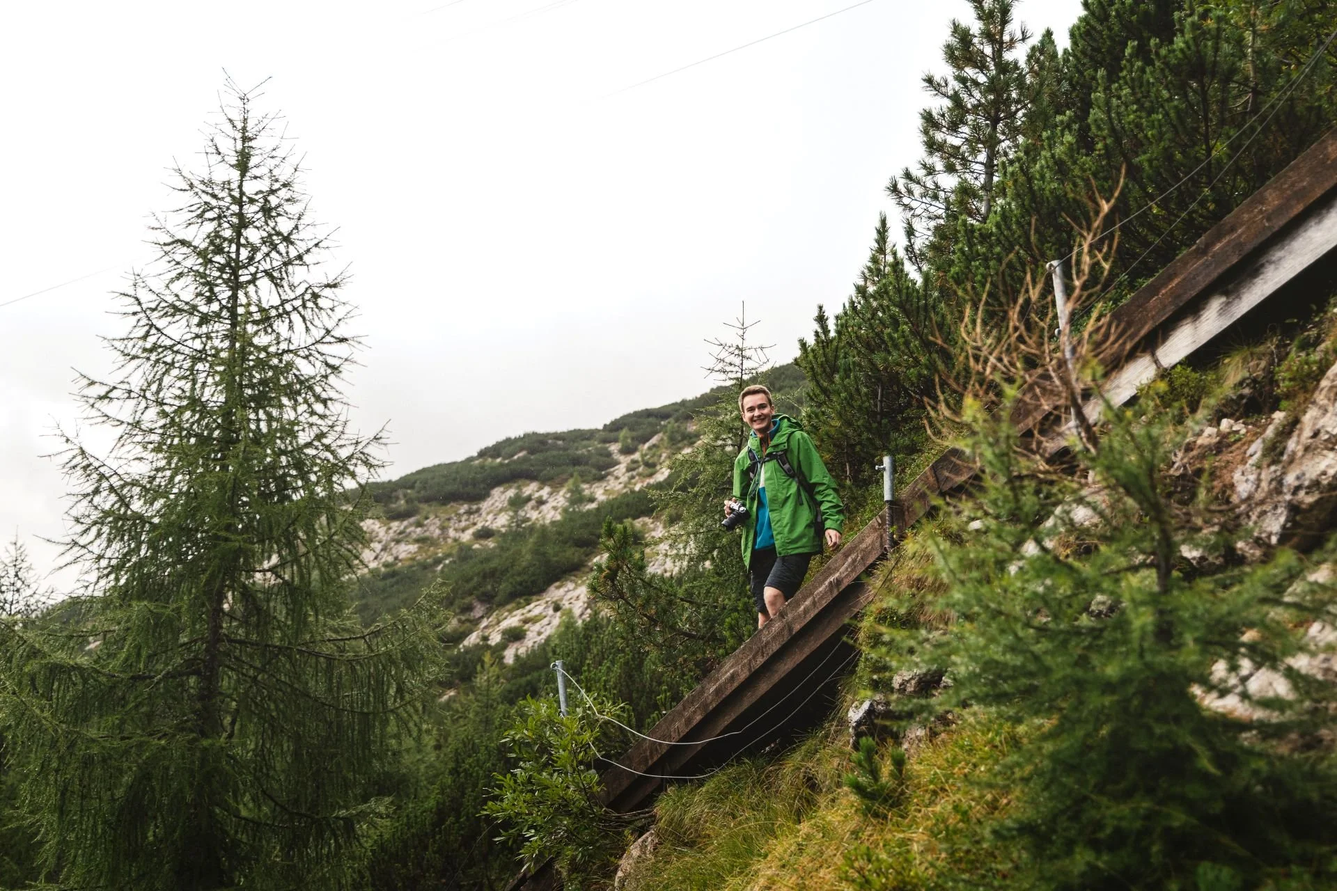 A person wearing a green jacket and black shorts hiking along a steep rocky trail with pine trees and mountain scenery.