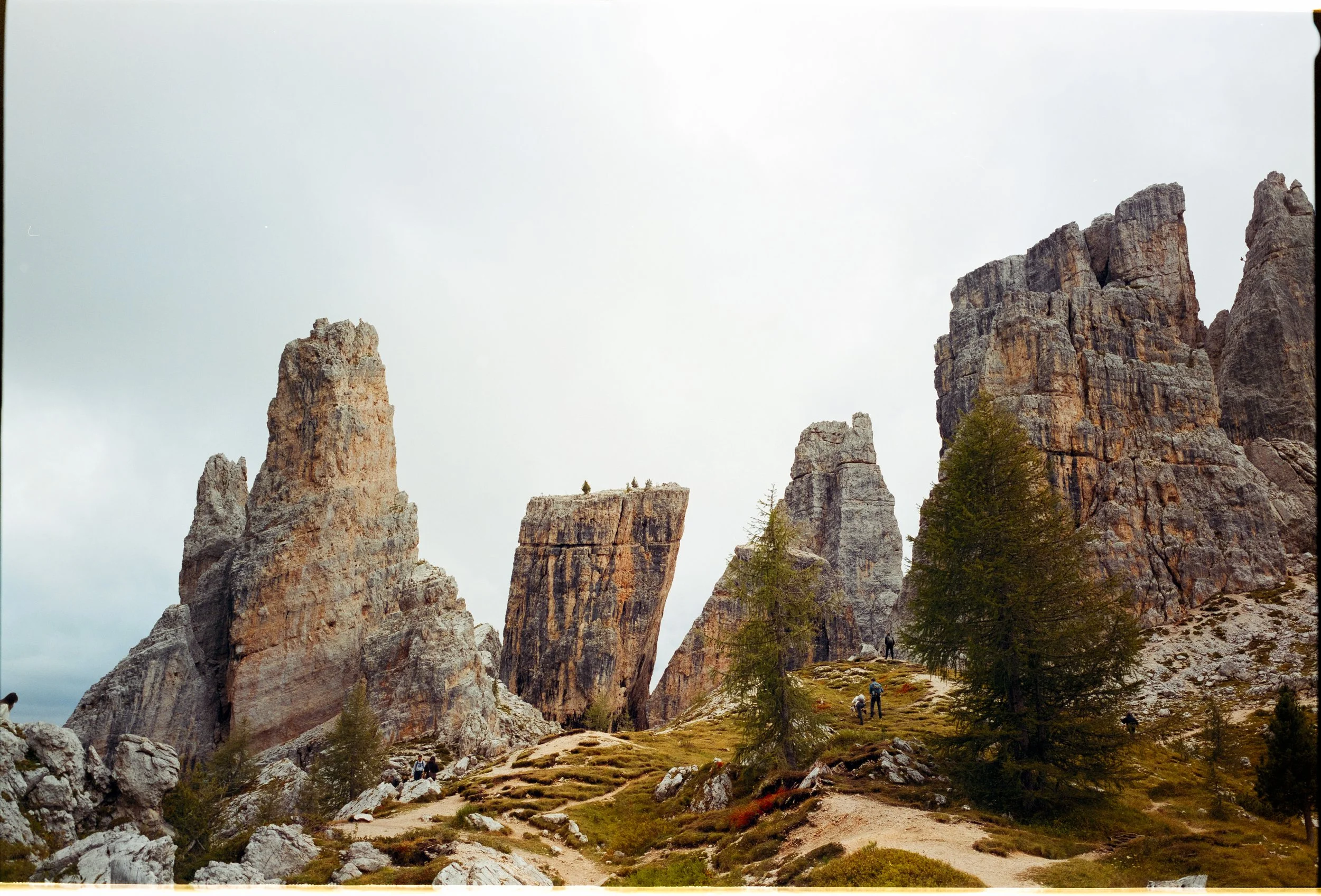 Tall rock formations in a mountainous landscape with hikers exploring the terrain, surrounded by trees and cloudy sky.
