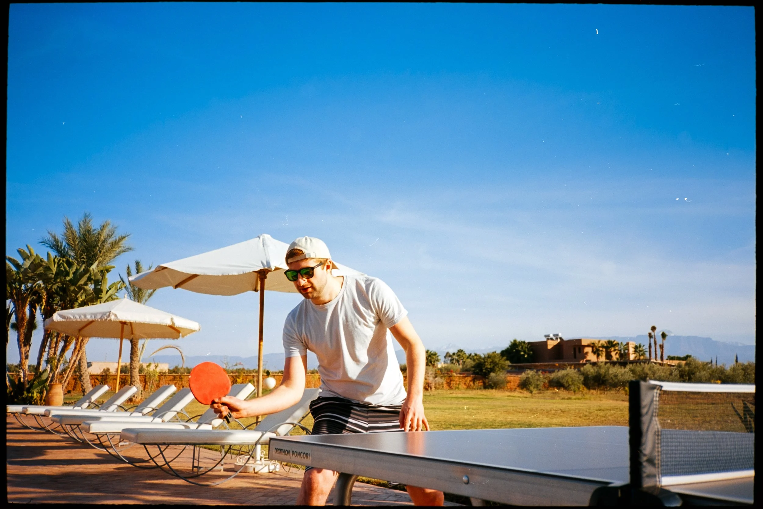 A young man playing ping pong outdoors under a bright blue sky with umbrellas and lounge chairs nearby, with palm trees and buildings in the background.
