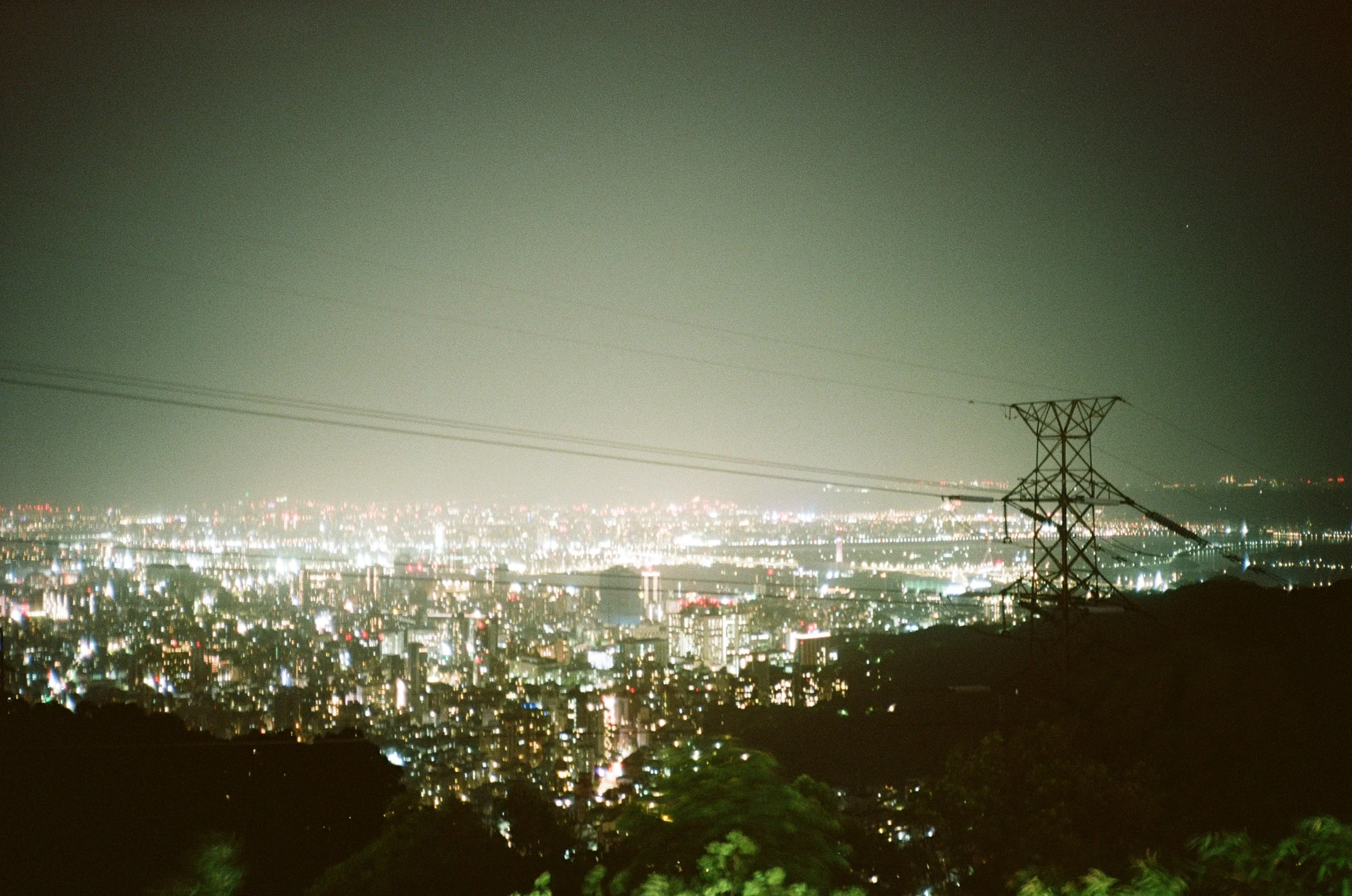 Nighttime cityscape of a densely built urban area illuminated with many lights, viewed from a hillside with an electricity pylon in the foreground and a hazy sky above.
