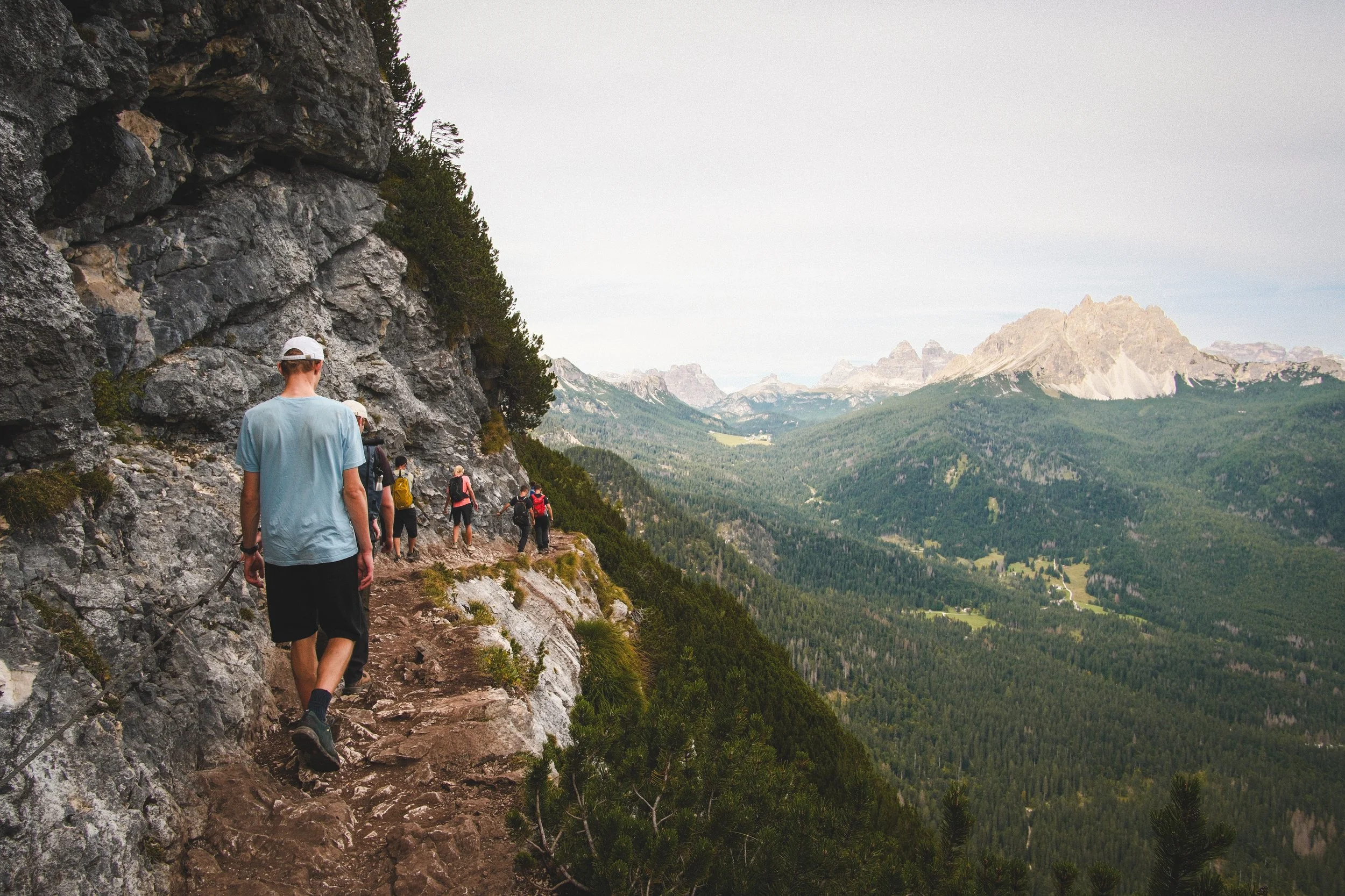 A group of hikers walking along a narrow mountain trail with a rocky cliff on the left and a vast green valley with mountains in the distance. The sky is overcast.