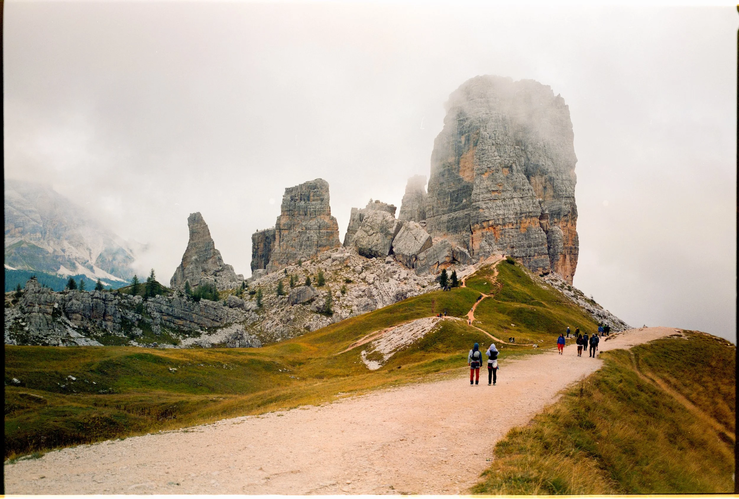 A group of hikers walking along a winding trail in a mountainous landscape, with large rock formations and peaks partly shrouded in mist in the background.