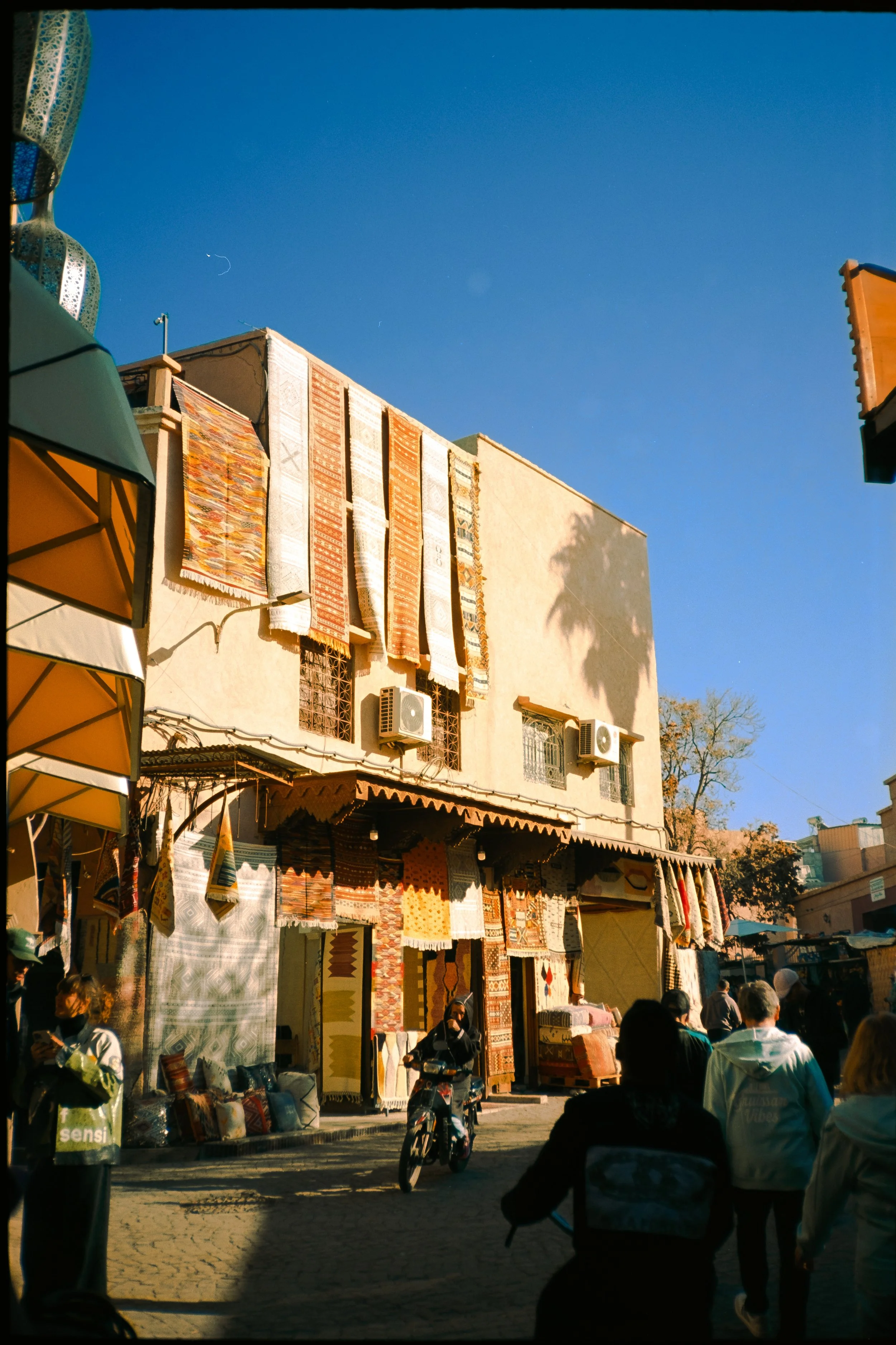 Outdoor market with fabric and rug stalls, people walking, and a building with hanging rugs and textiles, under a clear blue sky.