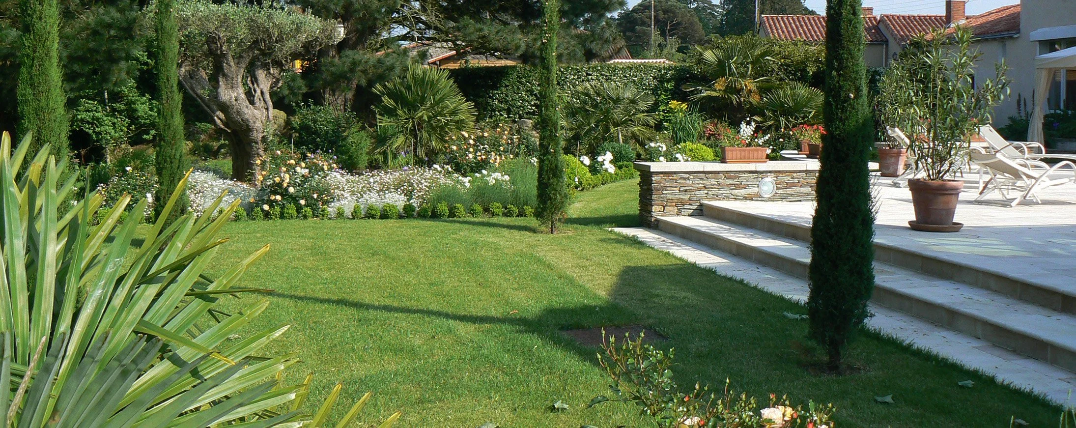 Jardin bien entretenu avec pelouse verte, divers arbres, buissons, fleurs colorées et terrasse avec chaises longues en plastique blanc.