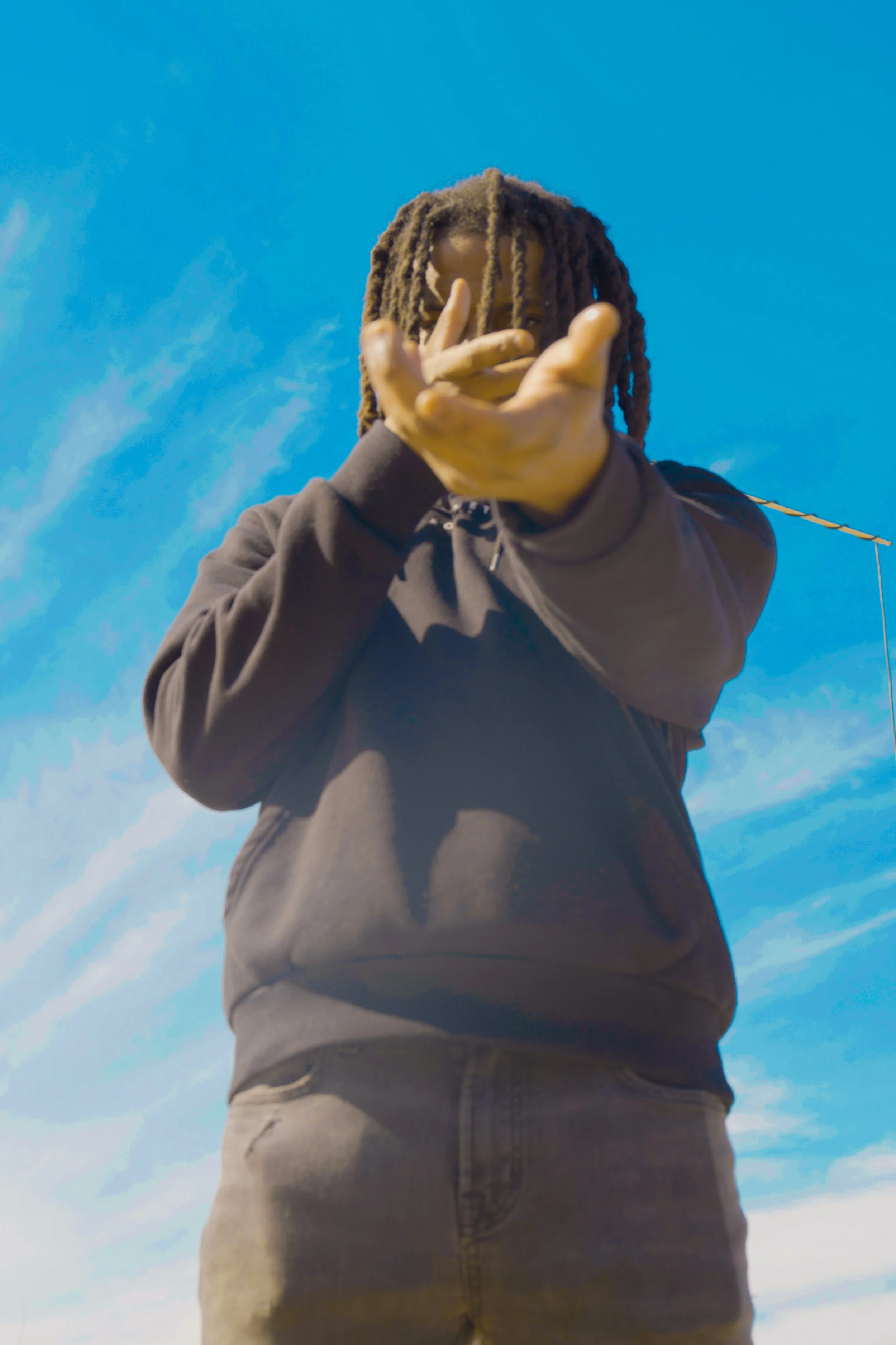 A person with dreadlocks wearing a black hoodie and beige pants, extending their hand toward the camera against a blue sky with some clouds.